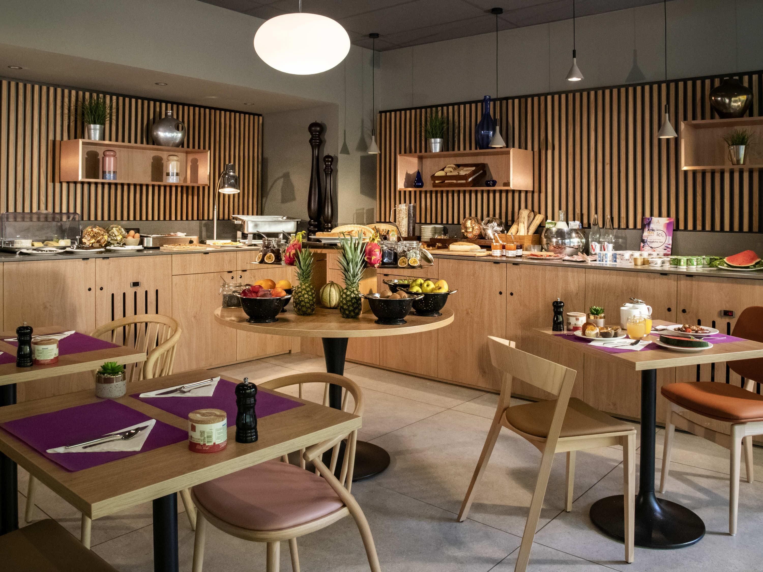A modern kitchen with wooden cabinets, a central island with fruits and beverages, and a dining area with chairs and tables.