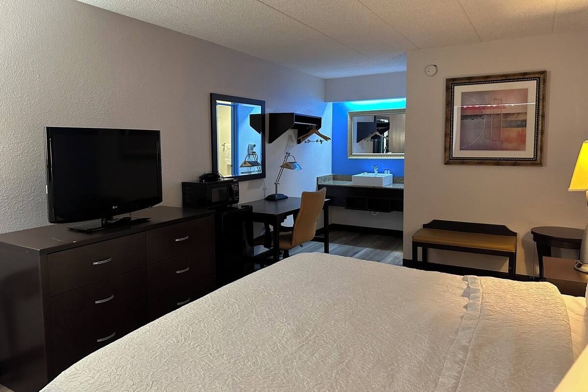 A hotel room with a bed, a desk with a computer, a television, a mirror, a sink, and a framed picture on the wall.
