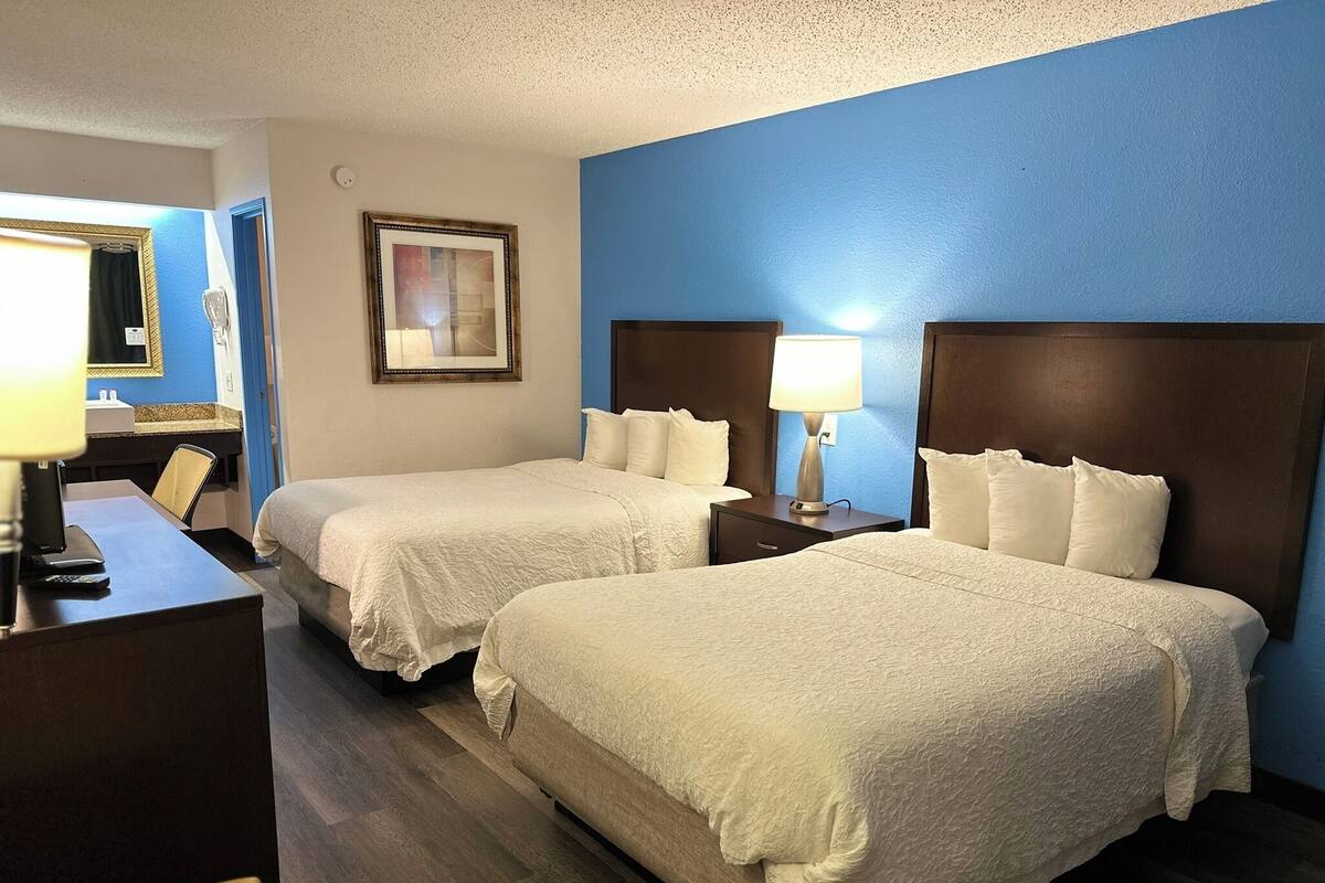 A hotel room with two beds, a desk, a chair, a lamp, a framed picture, and a blue wall.