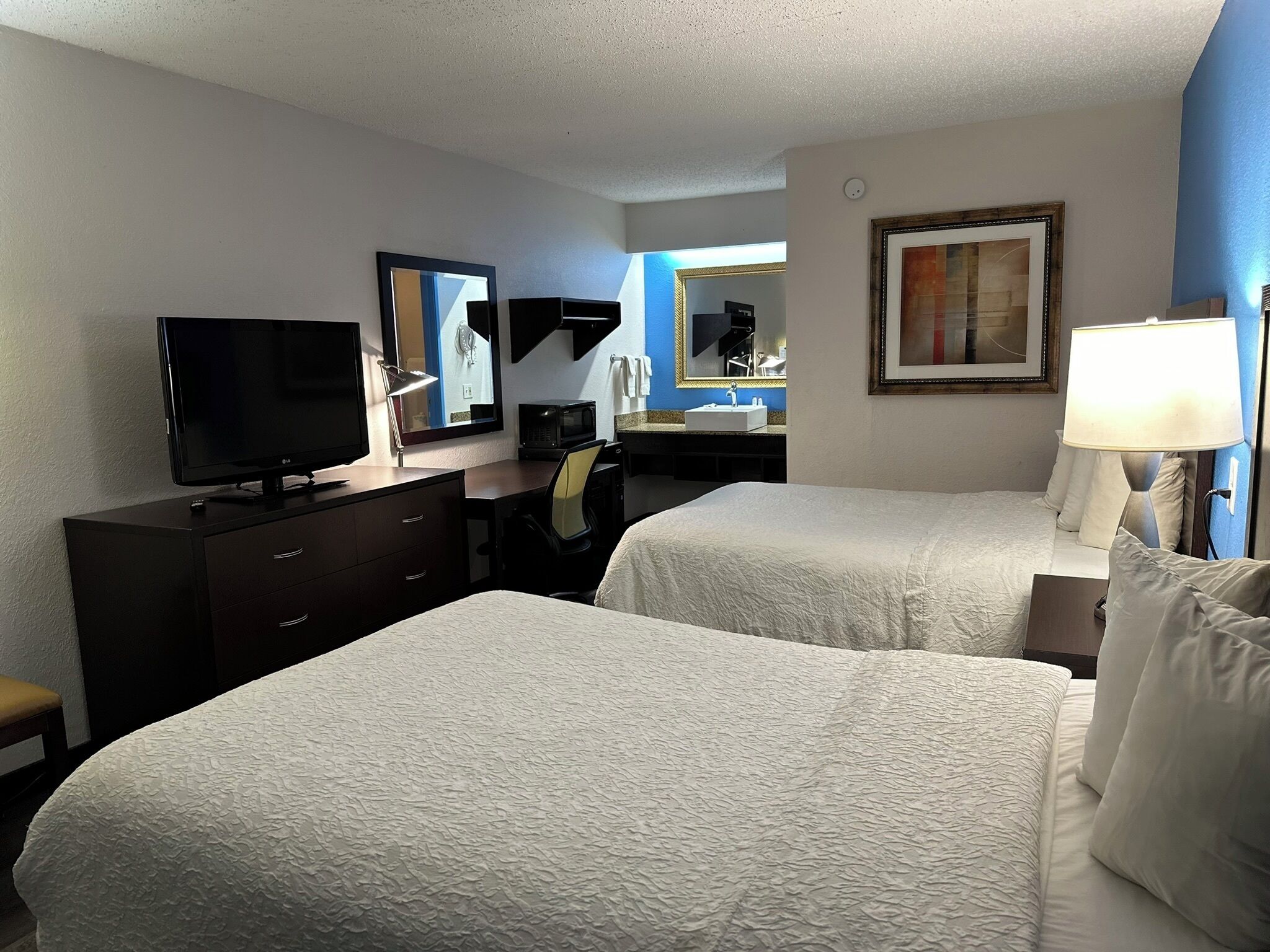 Una habitación de hotel con dos camas, un escritorio con computadora, televisión, espejo, lámpara y un cuadro en la pared.