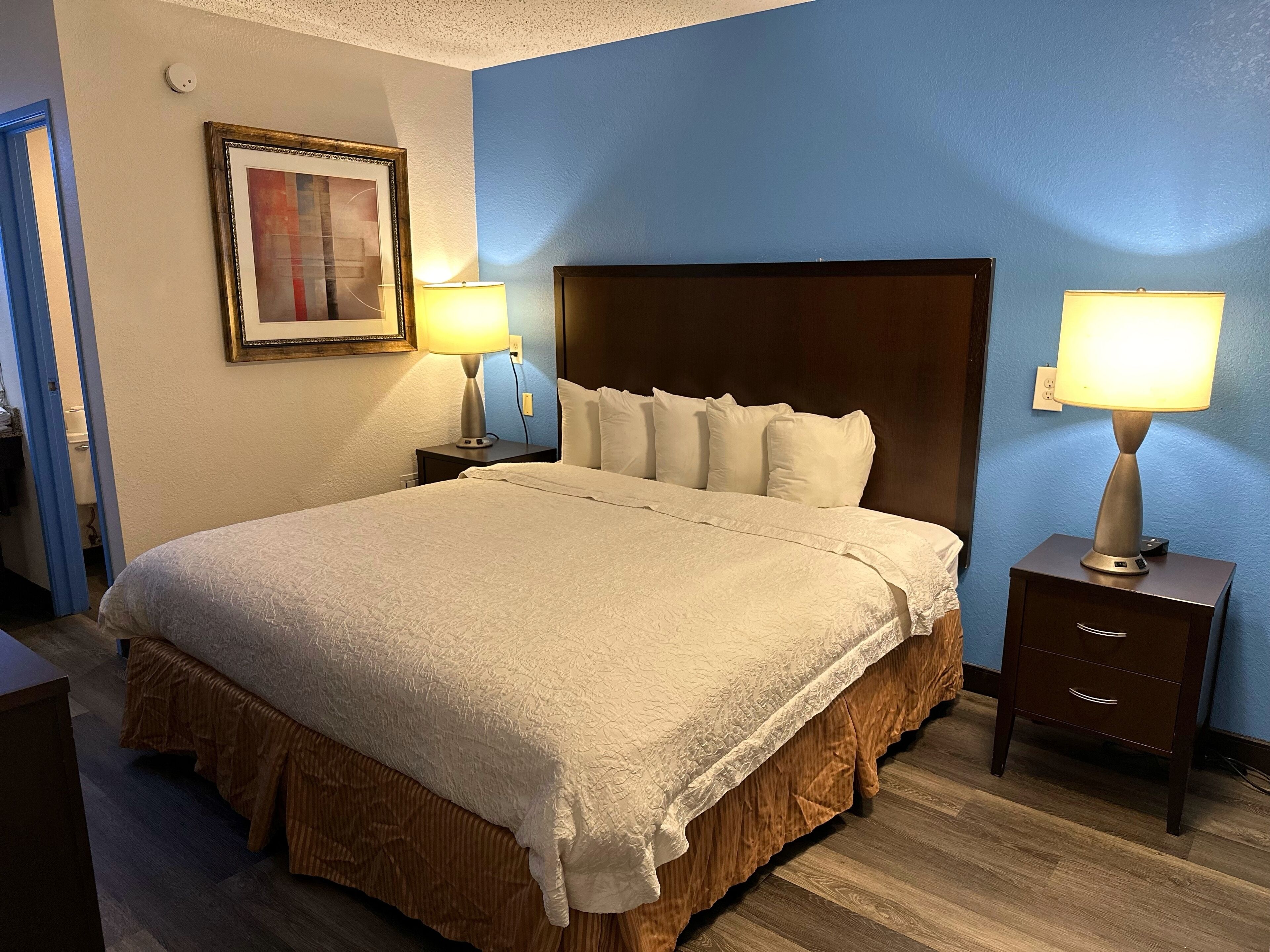 A hotel room with a large bed, two bedside tables, a nightstand, a framed picture on the wall, and a blue accent wall.
