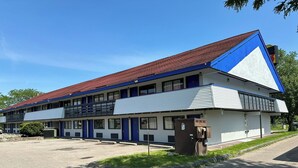 A two-story motel with a red roof and blue accents, featuring a parking lot and a small garden area.