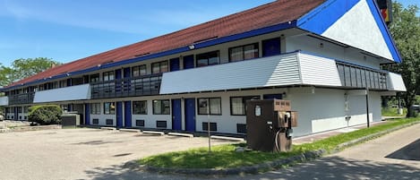 A two-story motel with a red roof and blue accents, featuring a parking lot and a small garden area.