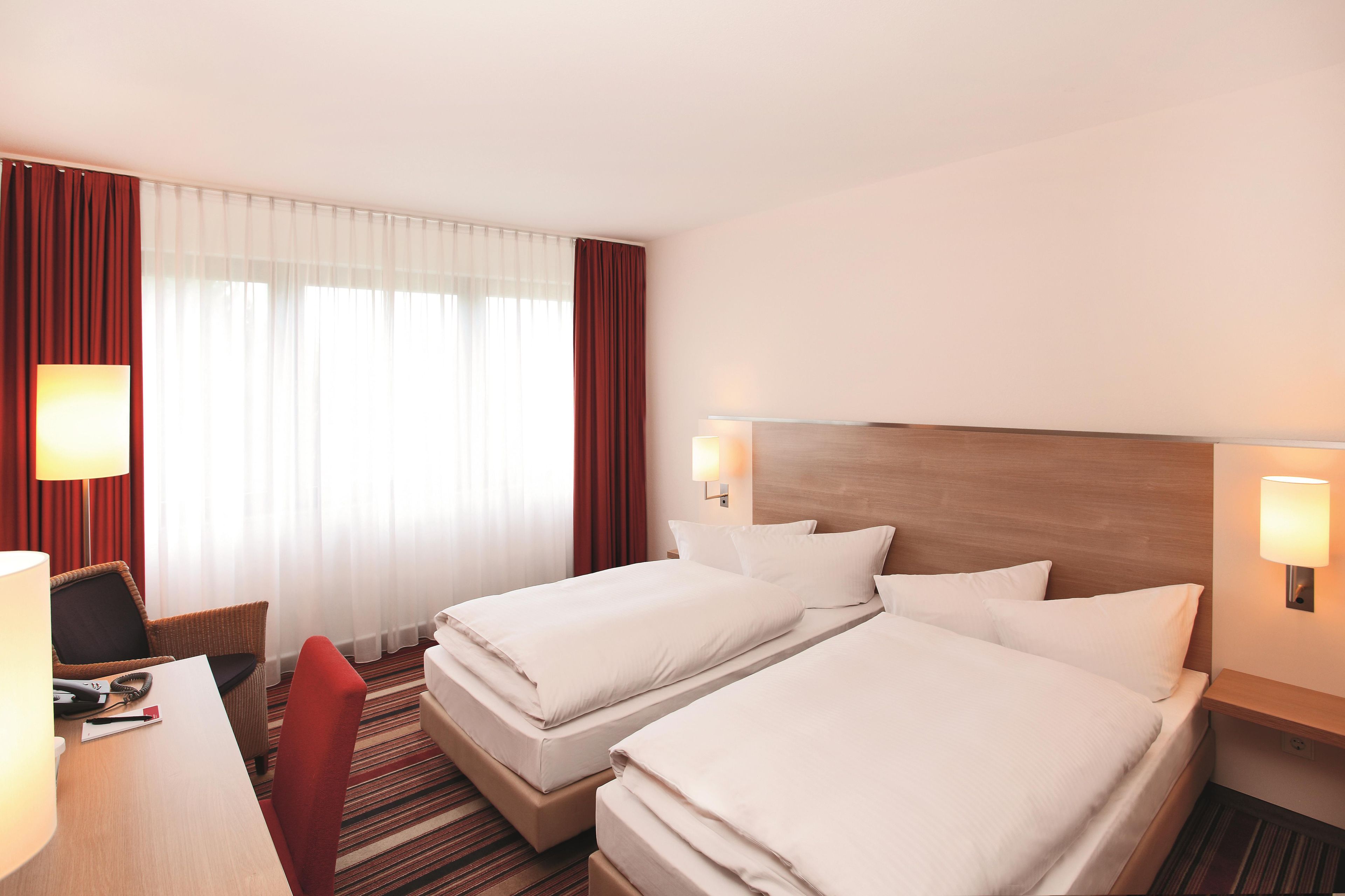 Ein Hotelzimmer mit zwei Betten, einem Schreibtisch, einem Stuhl und einem Fenster mit Vorhängen.