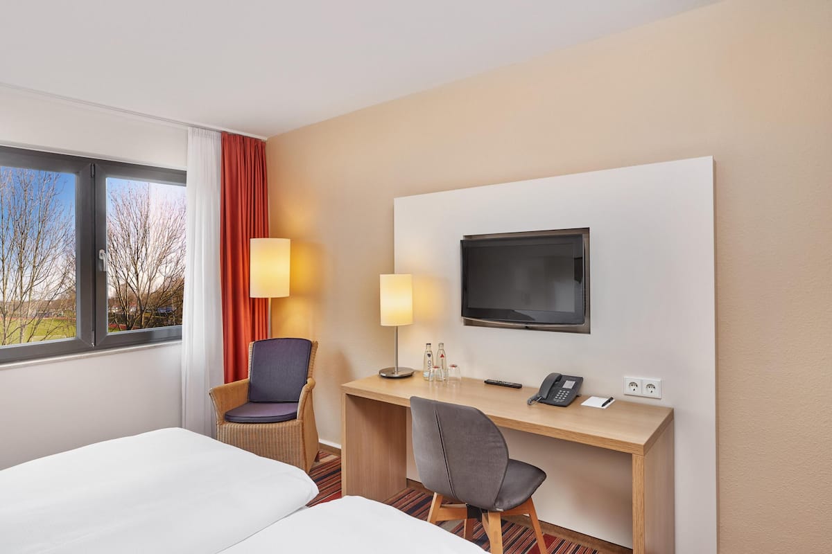 A hotel room with a bed, a desk with a TV, a chair, and a window with a view of trees.