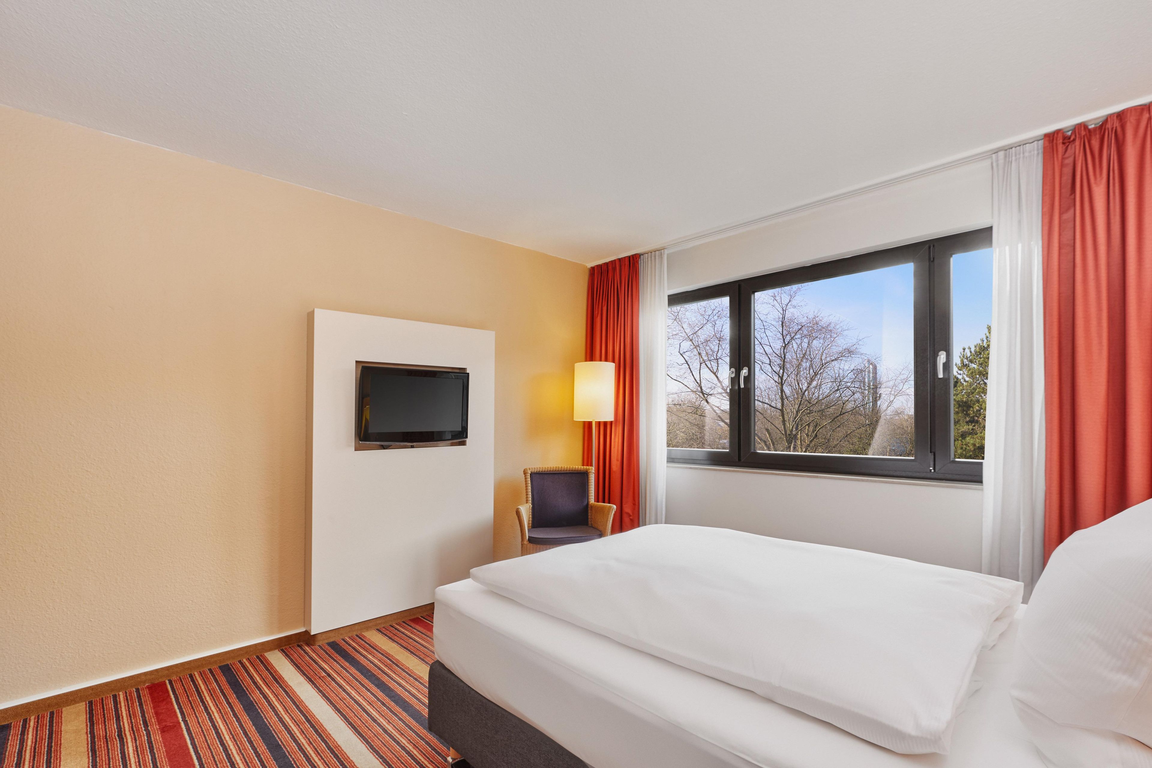 Ein Hotelzimmer mit Bett, Wandfernseher, Sessel und einem Fenster mit Vorhängen.