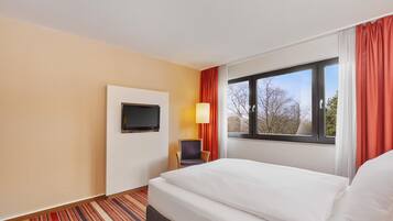 Ein Hotelzimmer mit Bett, Wandfernseher, Sessel und einem Fenster mit Vorhängen.
