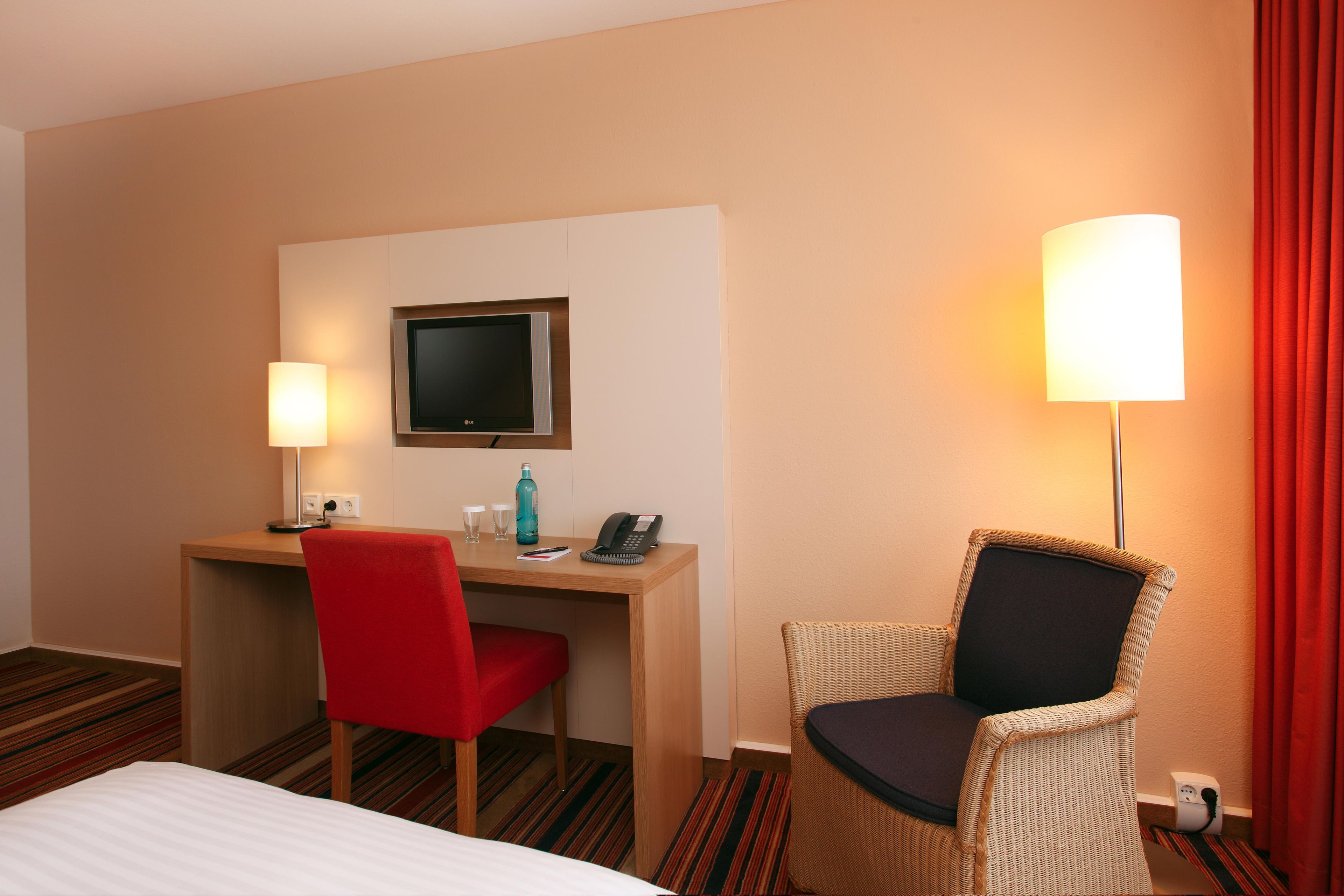 Ein Hotelzimmer mit einem Bett, einem Schreibtisch mit Flachbildfernseher, einem roten Sessel, einem Korbstuhl, einer Lampe und einem Telefon.