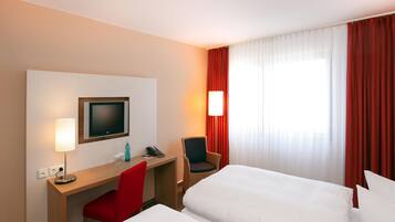 Ein Hotelzimmer mit einem Bett, einem Schreibtisch mit Fernseher, einem Sessel, einer Lampe und einem Vorhangfenster.