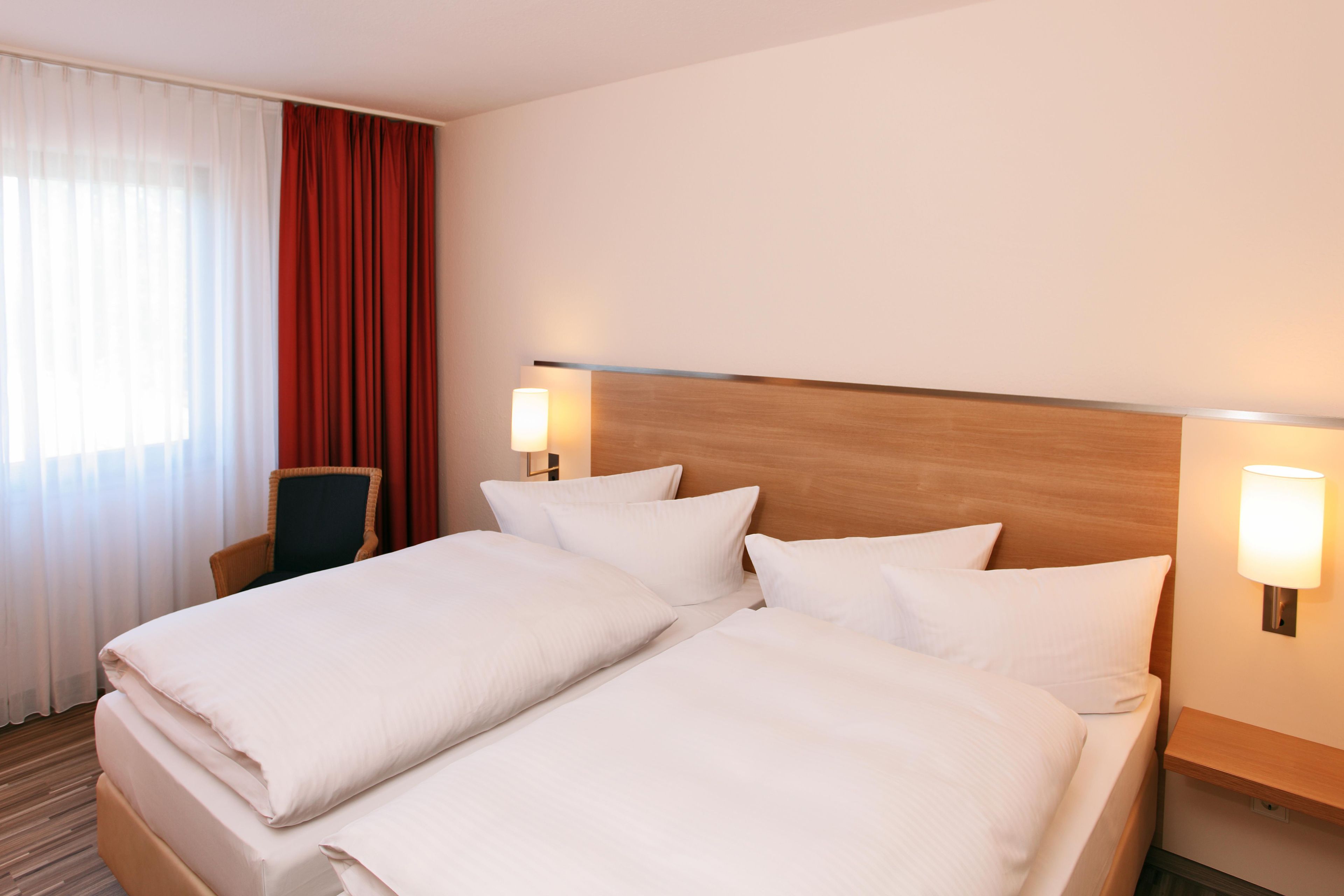 Una habitación de hotel con una cama doble, dos lámparas de noche, una silla y un ventanal con cortinas.