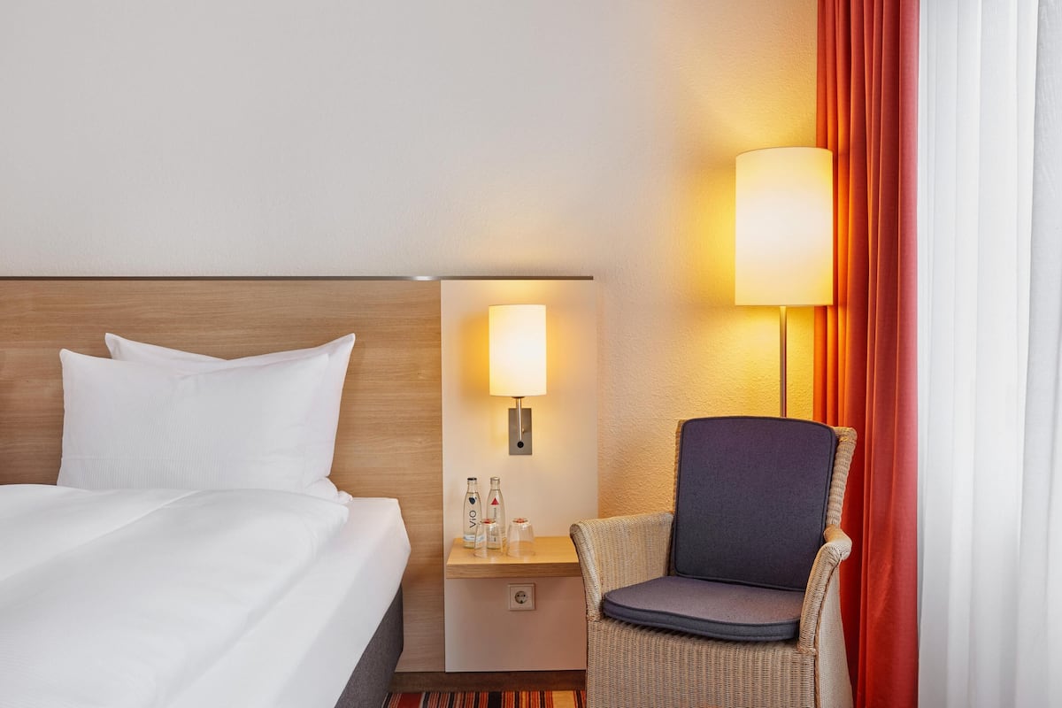 A hotel room with a bed, a nightstand with a lamp, a chair, and a window with curtains.