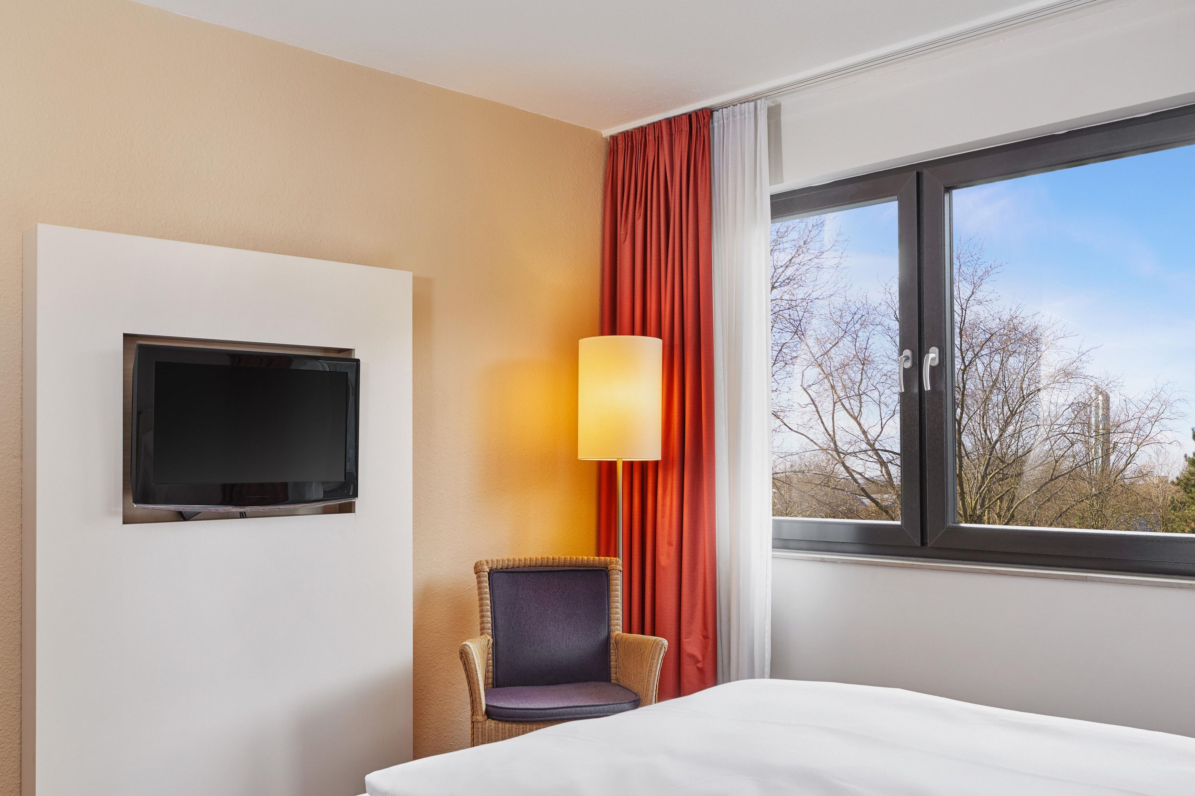 Ein Hotelzimmer mit Bett, Wandfernseher, Stuhl, Vorhängen an einem Fenster und Lampe.