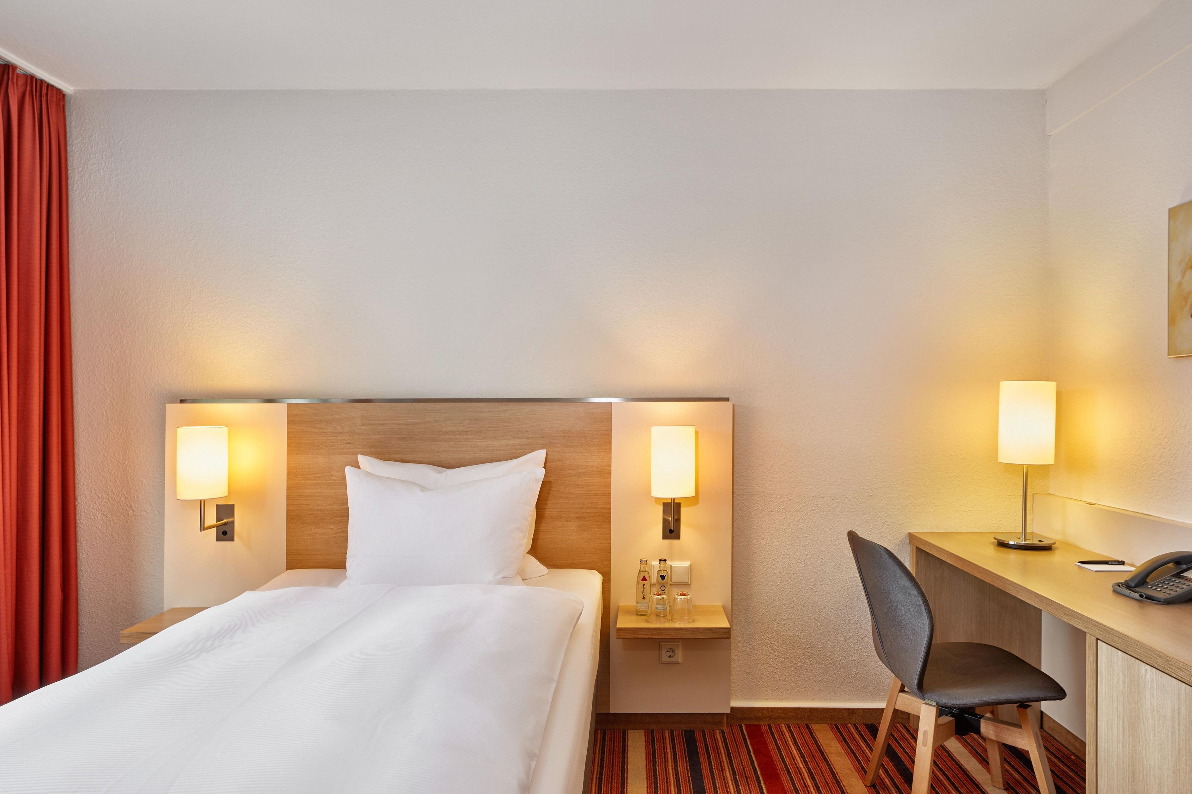 Ein Hotelzimmer mit einem Bett, zwei Nachttischlampen, einem Schreibtisch mit Stuhl und einem Telefon.