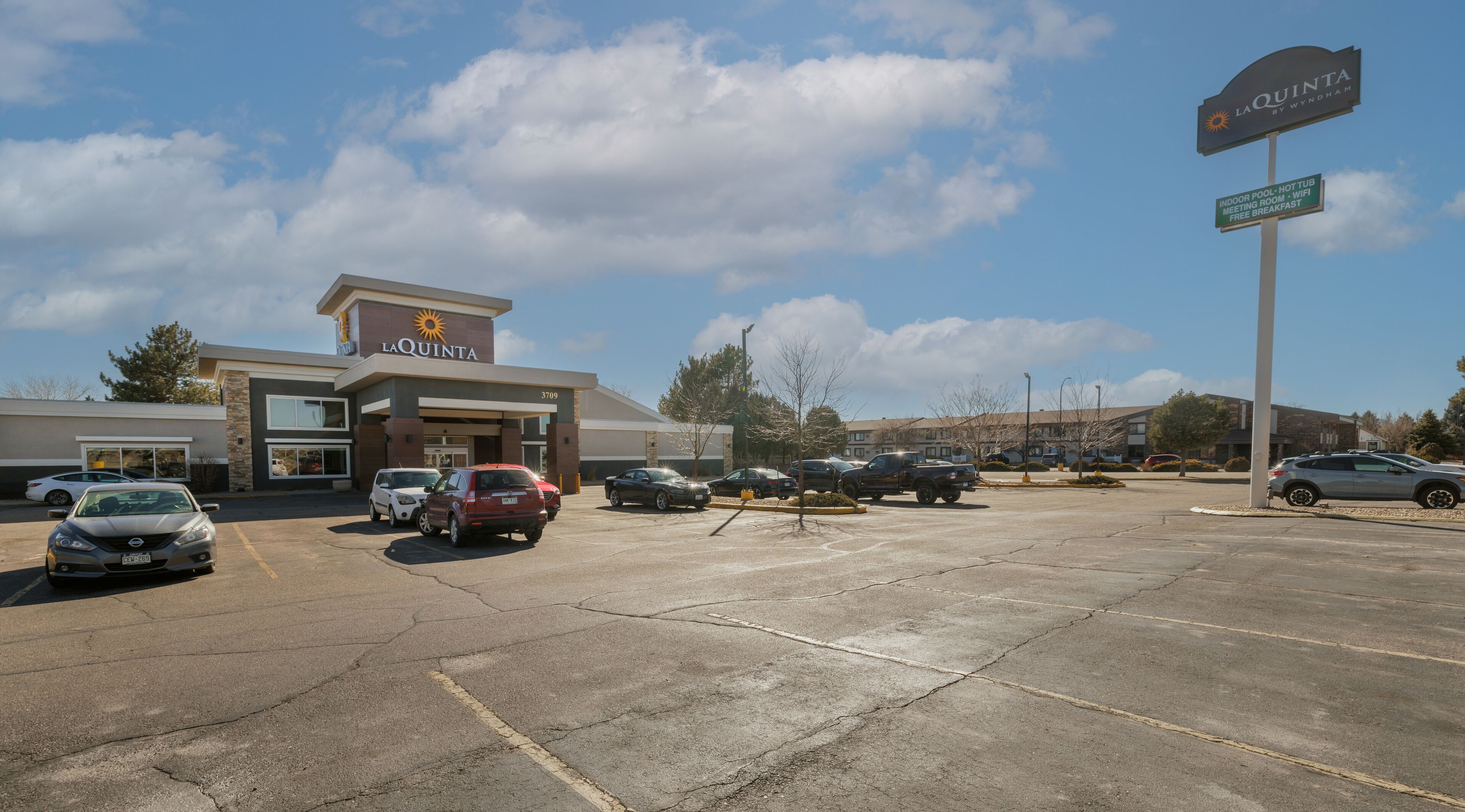 A parking lot with several cars, a La Quinta hotel, and a sign indicating amenities like an indoor pool, hot tub, meeting room, WiFi, and free breakfast.
