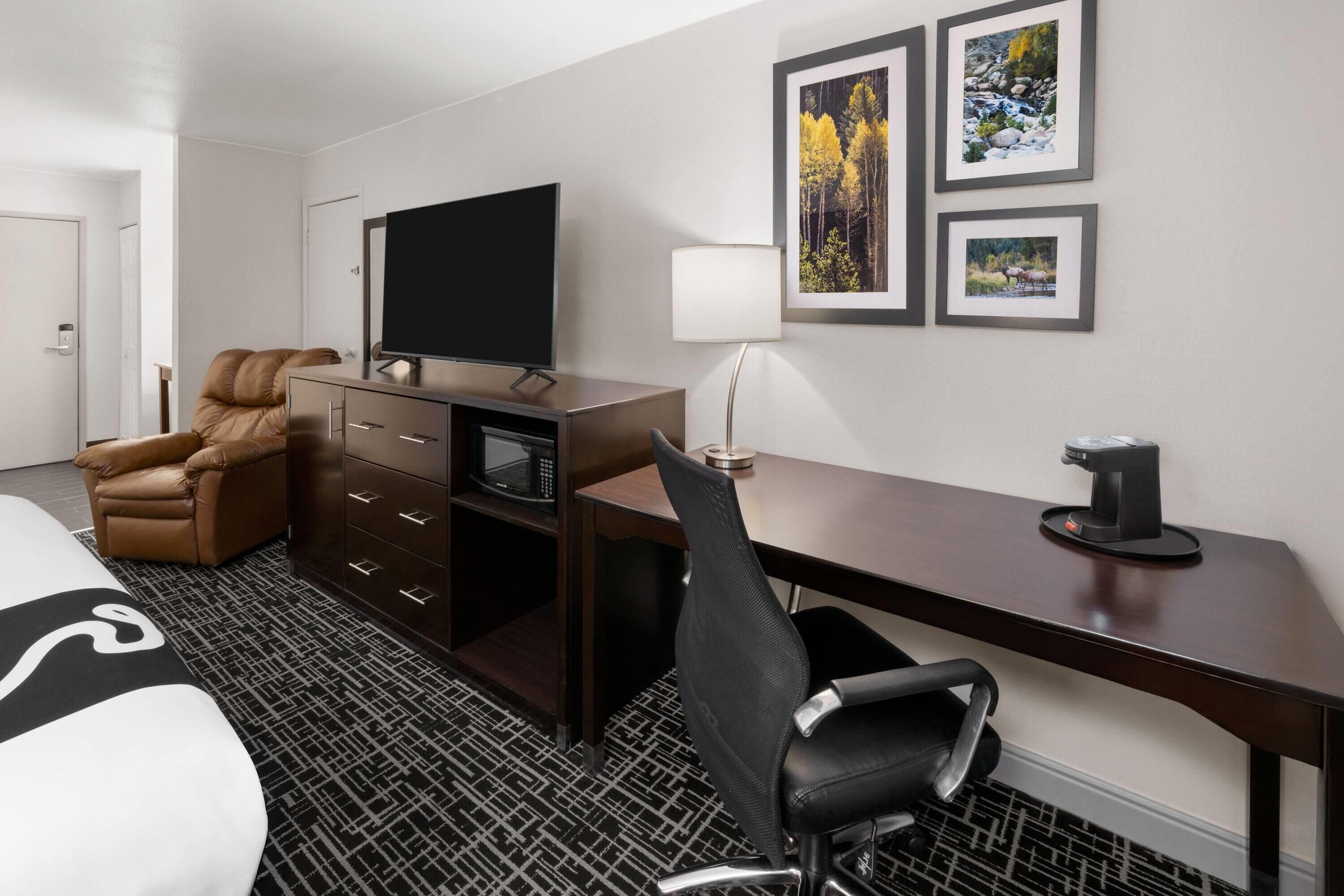 Habitación de hotel con cama, escritorio con computadora, televisor, silla, cafetera y cuadros en la pared.