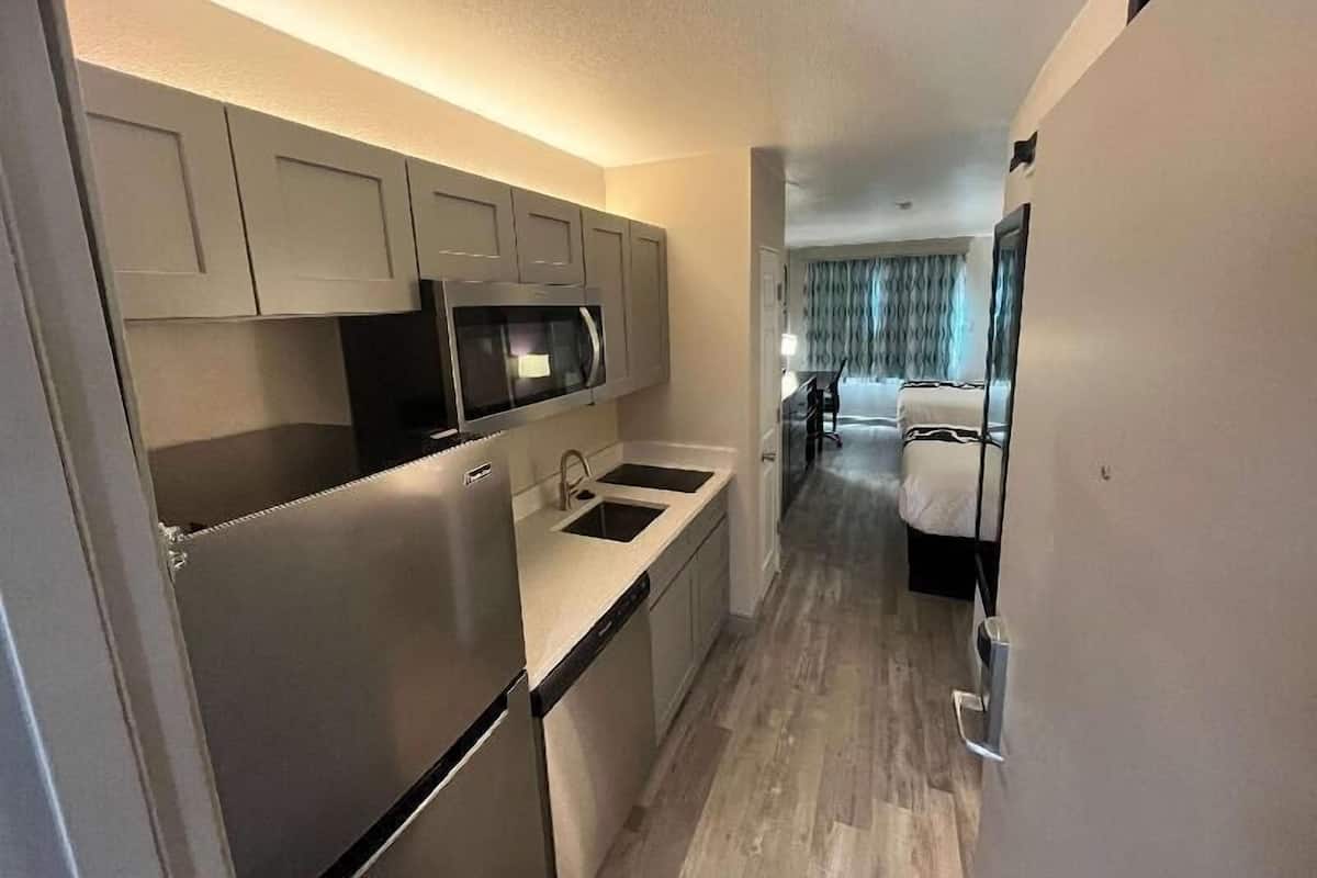 A compact kitchen with a refrigerator, microwave, and sink, leading to a bedroom with a bed and a chair.