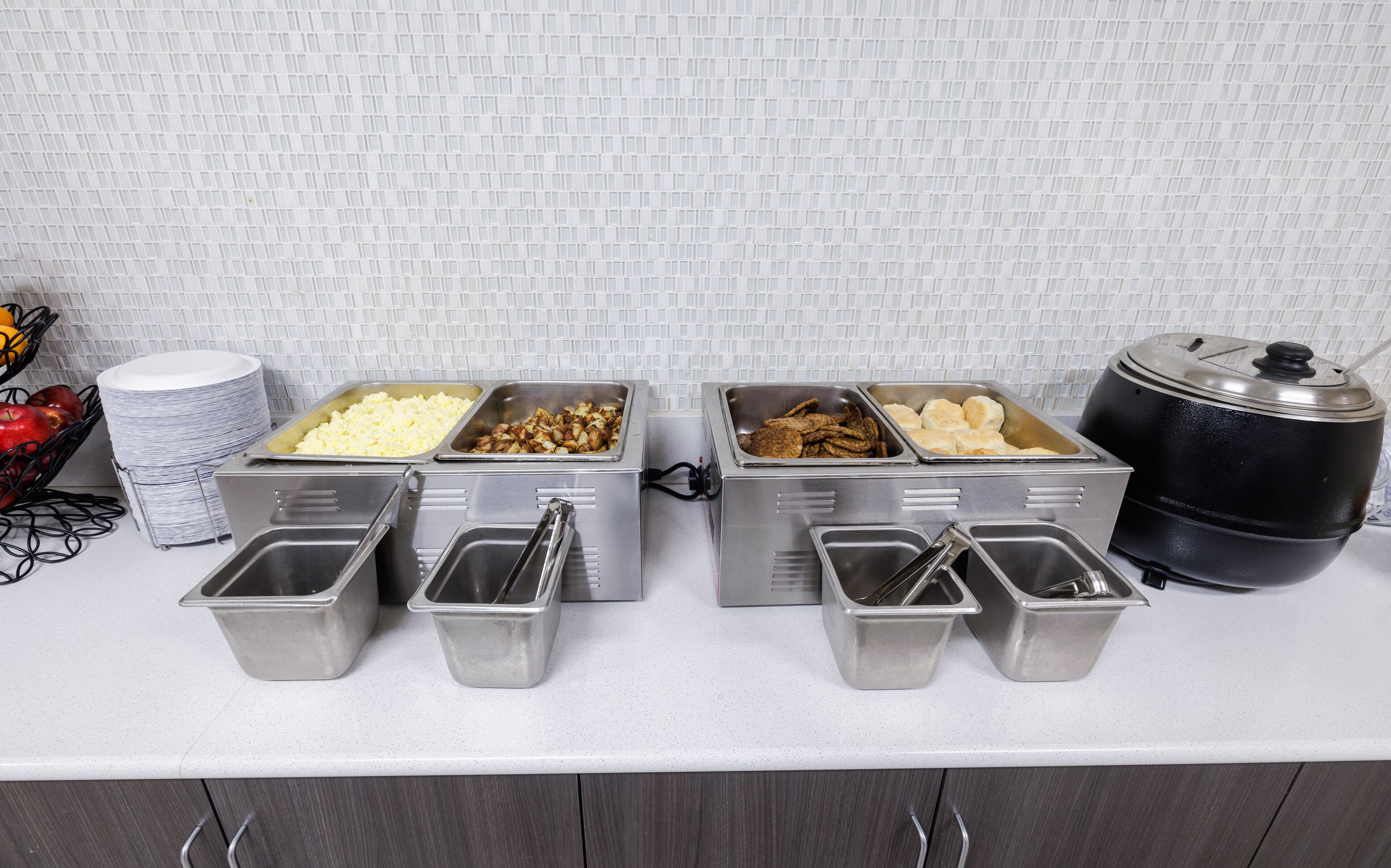 A buffet setup with various food items in metal containers, a stack of white plates, and a black pot on the counter.