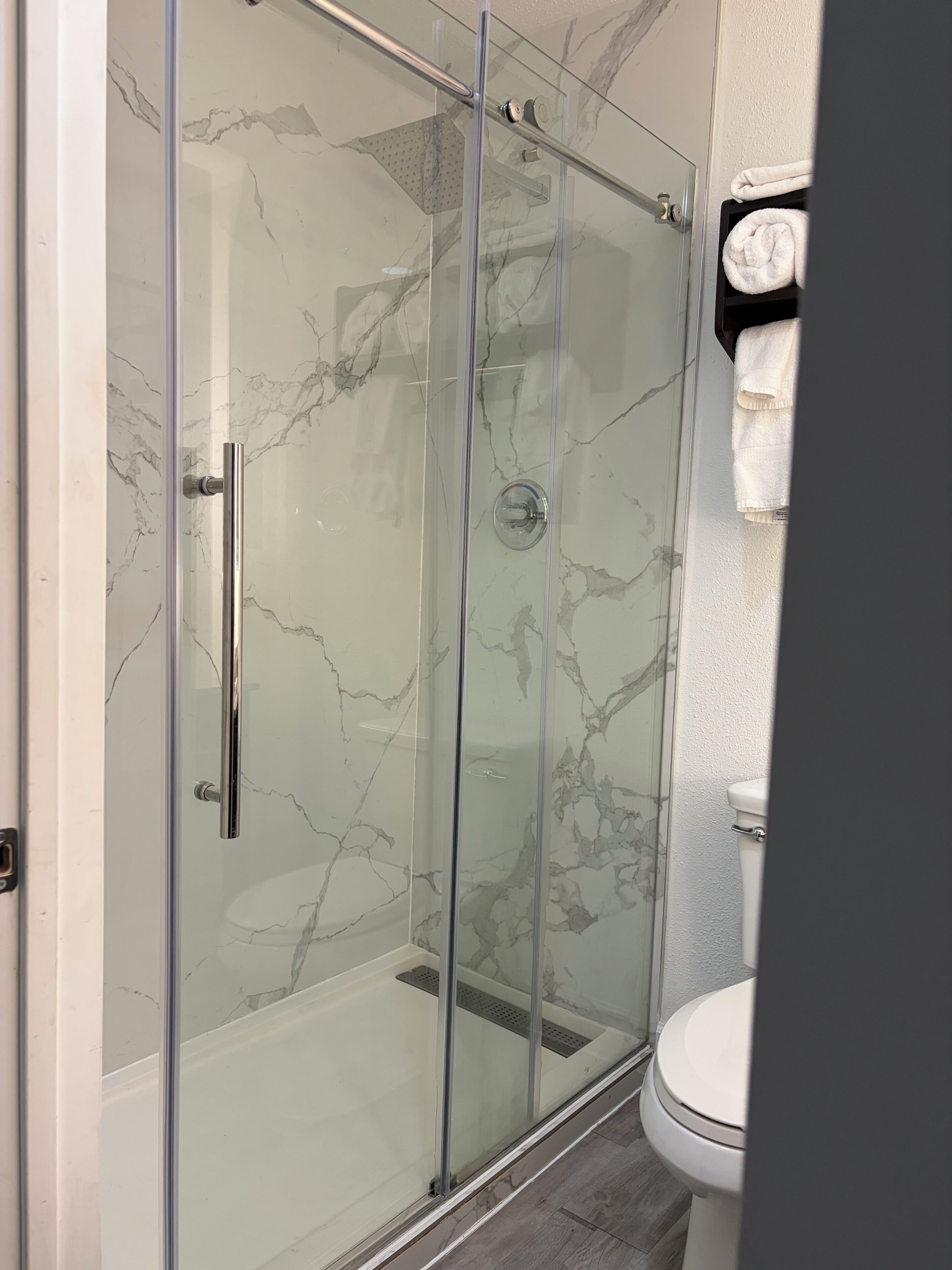 En brusekabine med glasvægge, et vægdesign der ligner marmor, et hvidt toilet og et håndklædetørrestativ med foldede håndklæder.