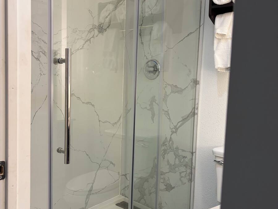 Ducha con cerramiento de vidrio, diseño de pared tipo mármol, inodoro blanco y un perchero con toallas dobladas.