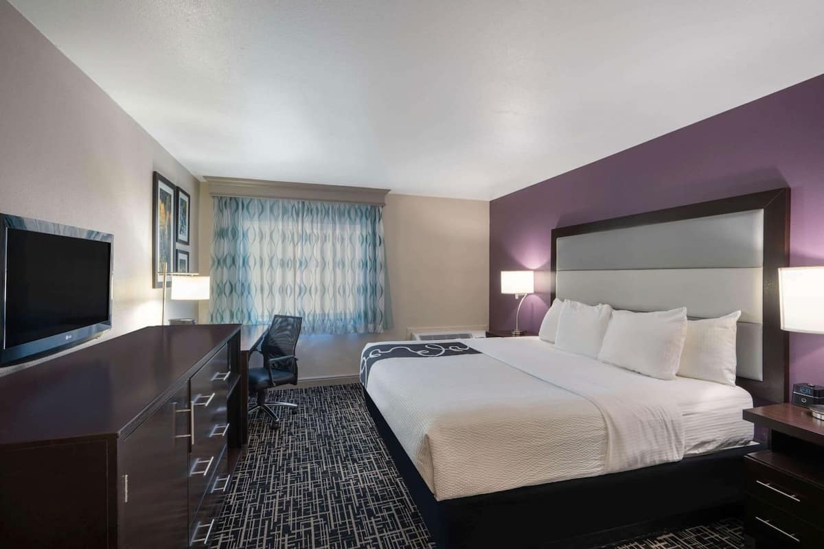 A hotel room with a bed, a desk with a TV, a chair, a window with curtains, and a nightstand with a lamp.