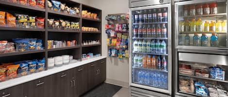 Zona de aperitivos y bebidas con estantes llenos de papas fritas y un refrigerador que contiene agua y refrescos.