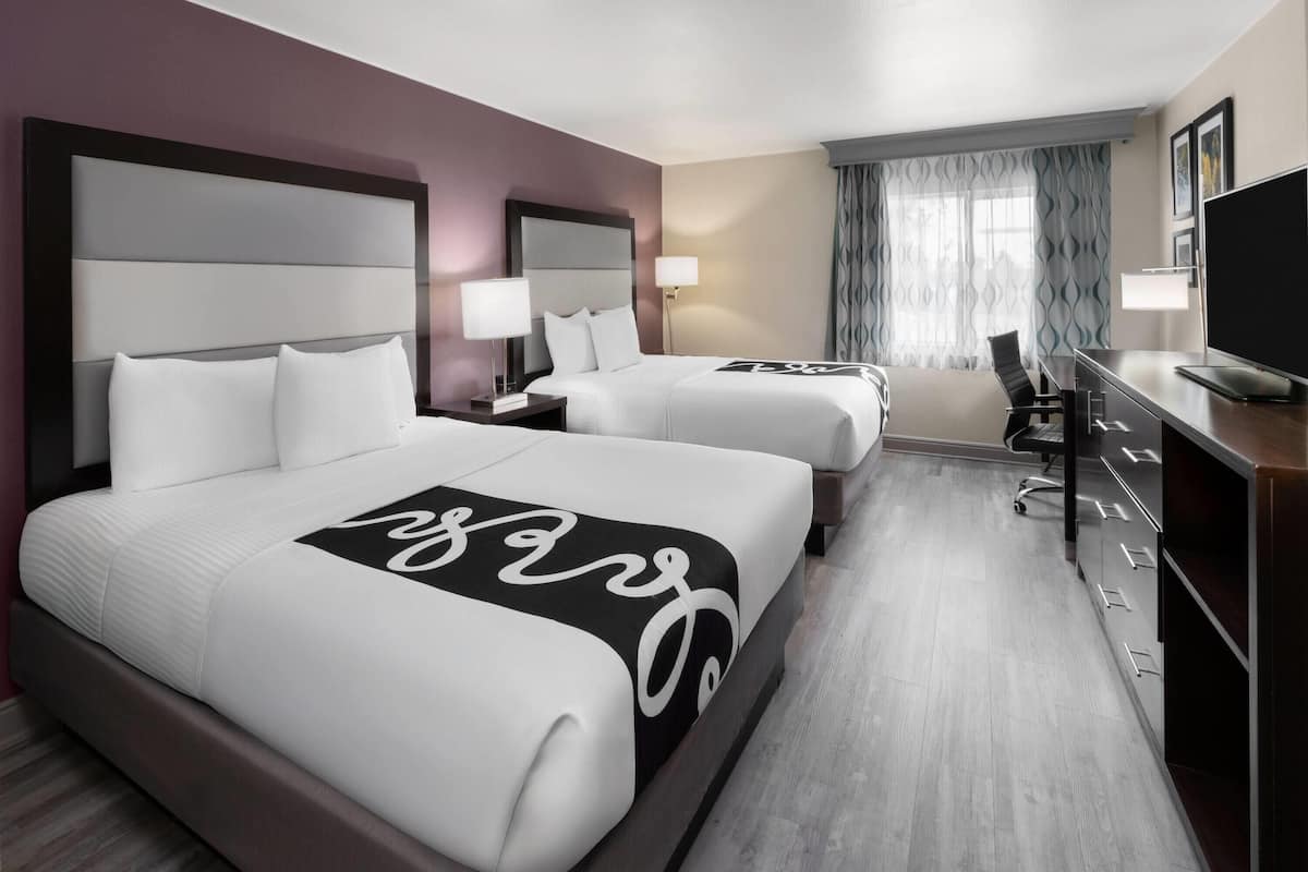 A hotel room with two beds, a desk, a chair, a TV, and a window with curtains.