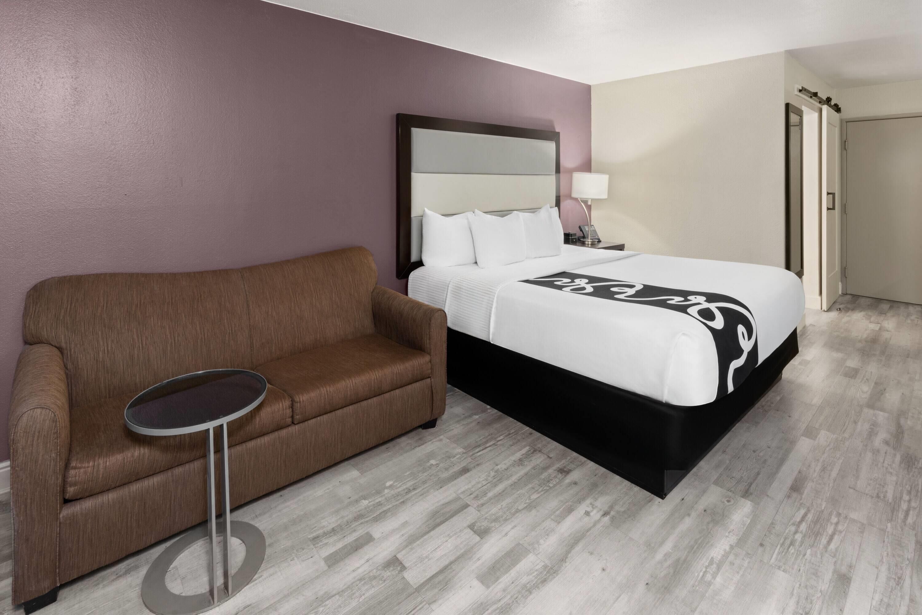 A hotel room with a bed, a brown sofa, a small round table, and a nightstand with a lamp.