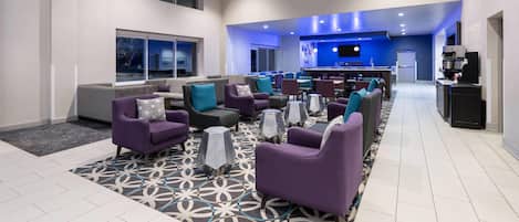 A modern hotel lobby with purple seating, a patterned rug, and a bar area.