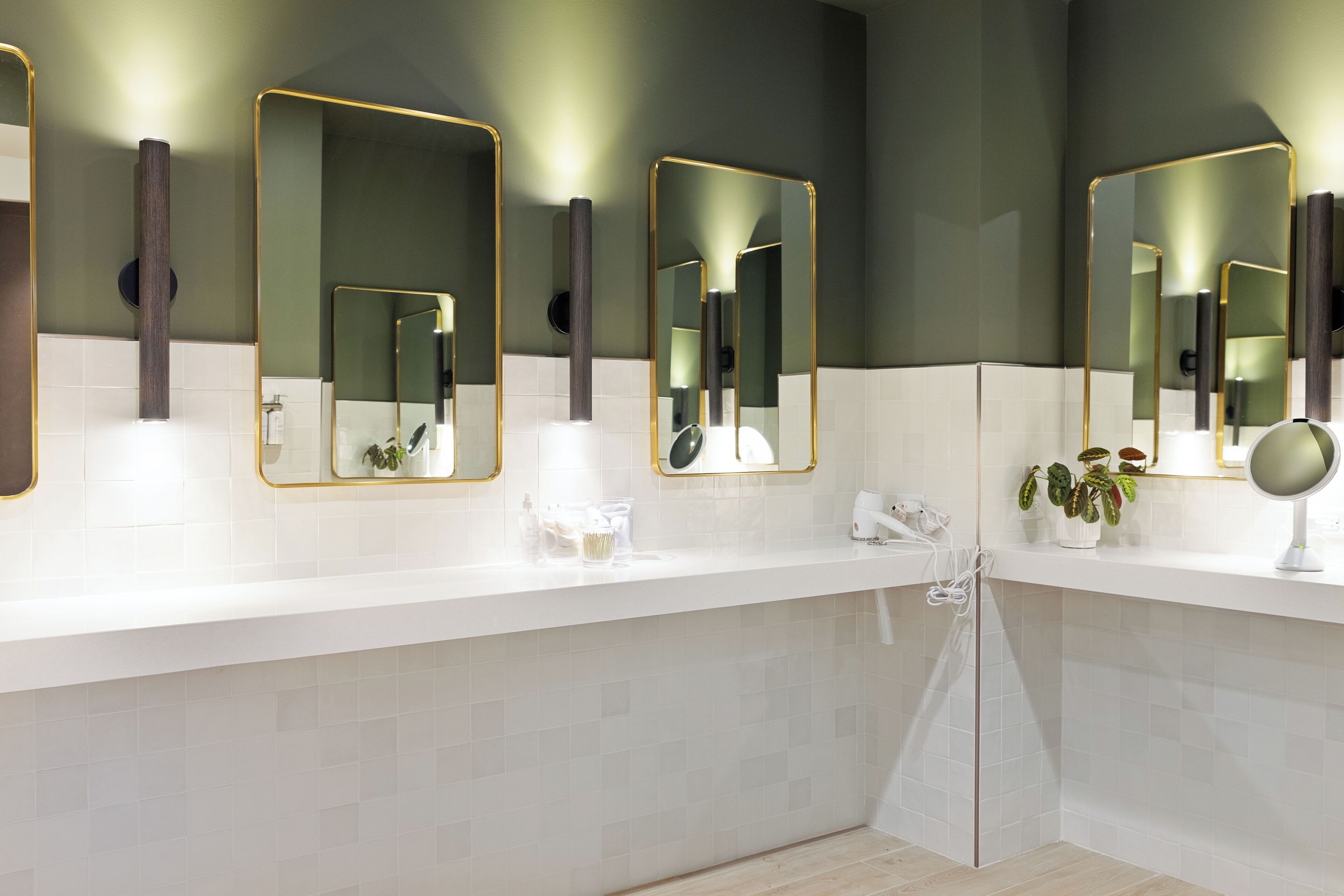 A modern bathroom with white tiled walls, a white countertop, and three mirrors with gold frames.