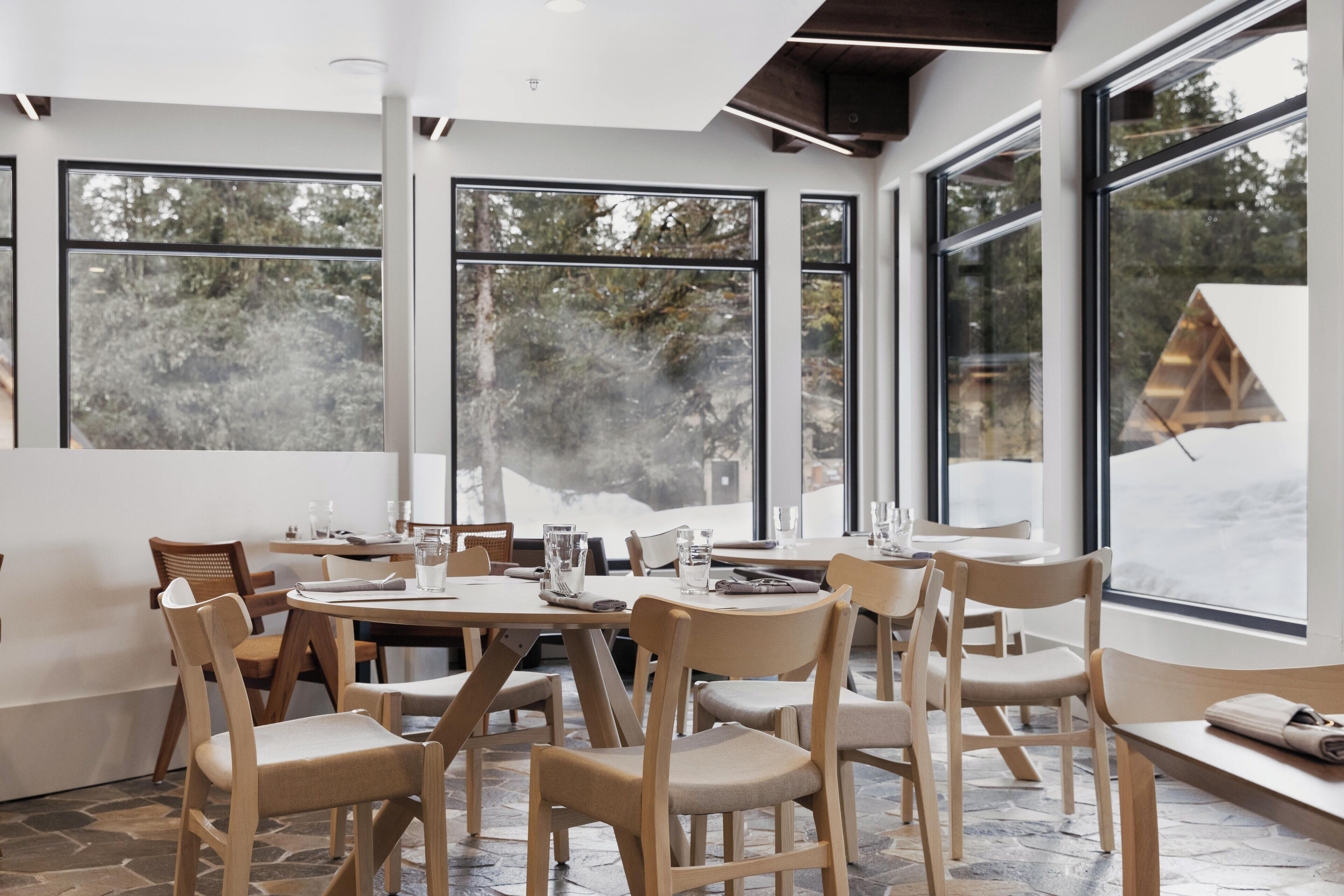 A modern dining area with large windows, wooden chairs, and a stone-tiled floor.