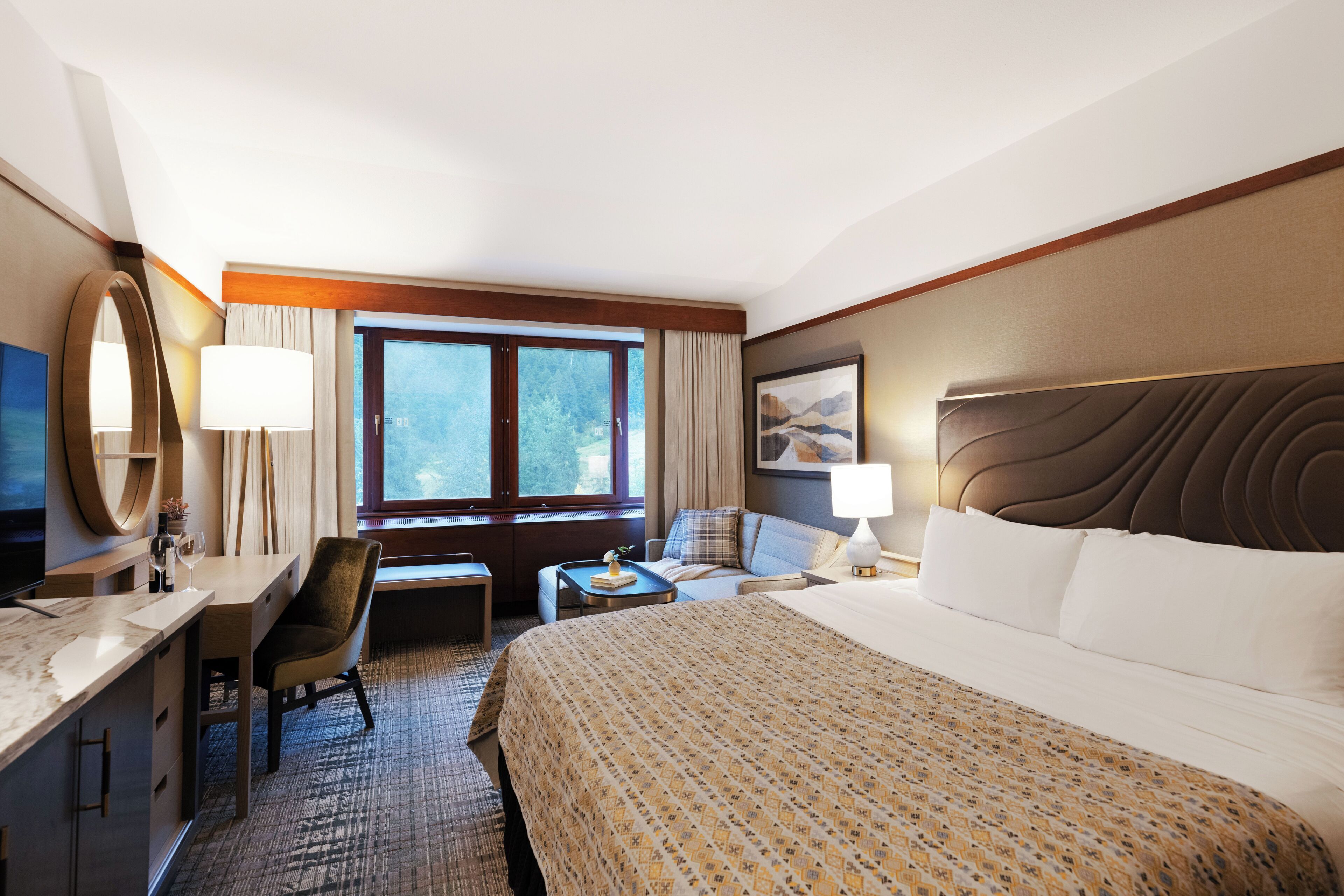 A hotel room with a large bed, a desk with a chair, a small table, a TV, and a window with curtains.