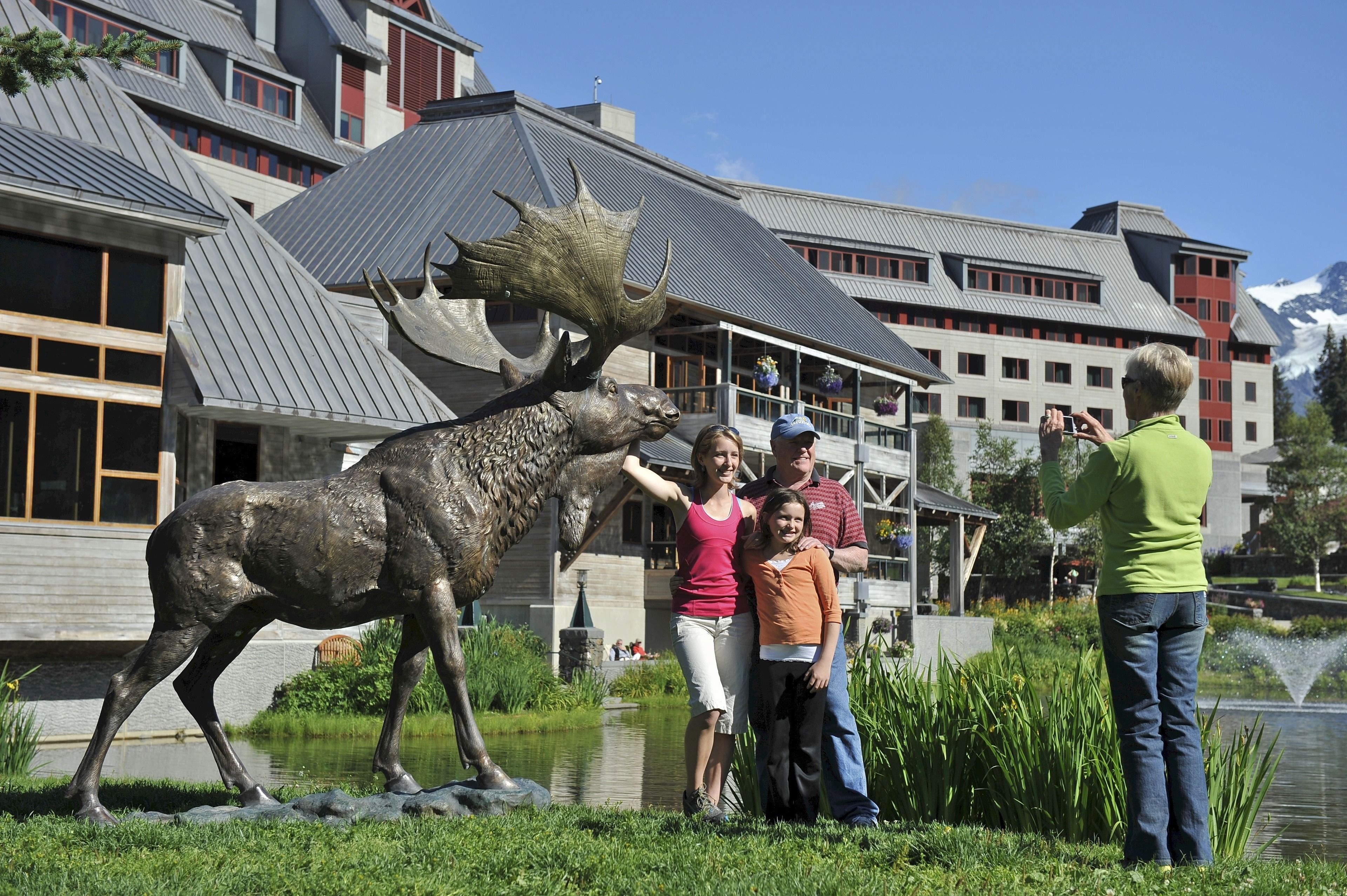 A family posing with a moose statue in front of a building with a sloped roof and a pond nearby.