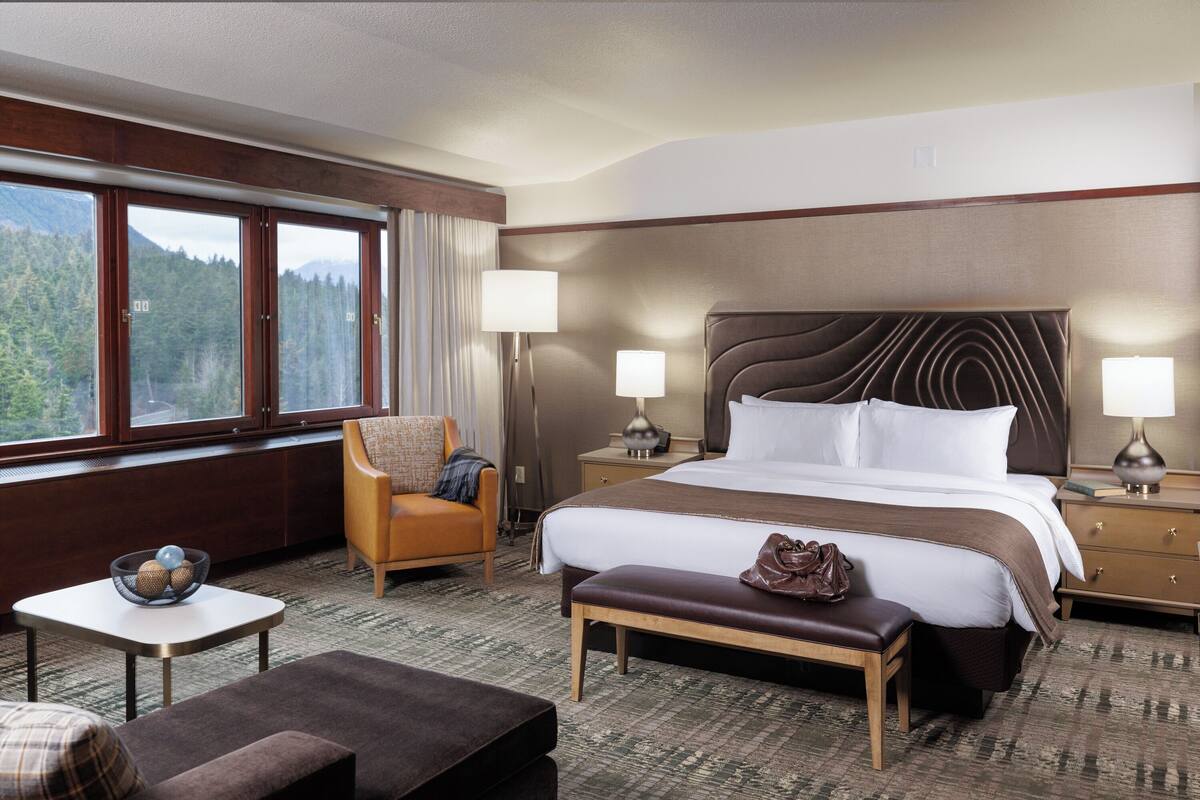 A hotel room with a large bed, a chair, a small table, and a view of the mountains.