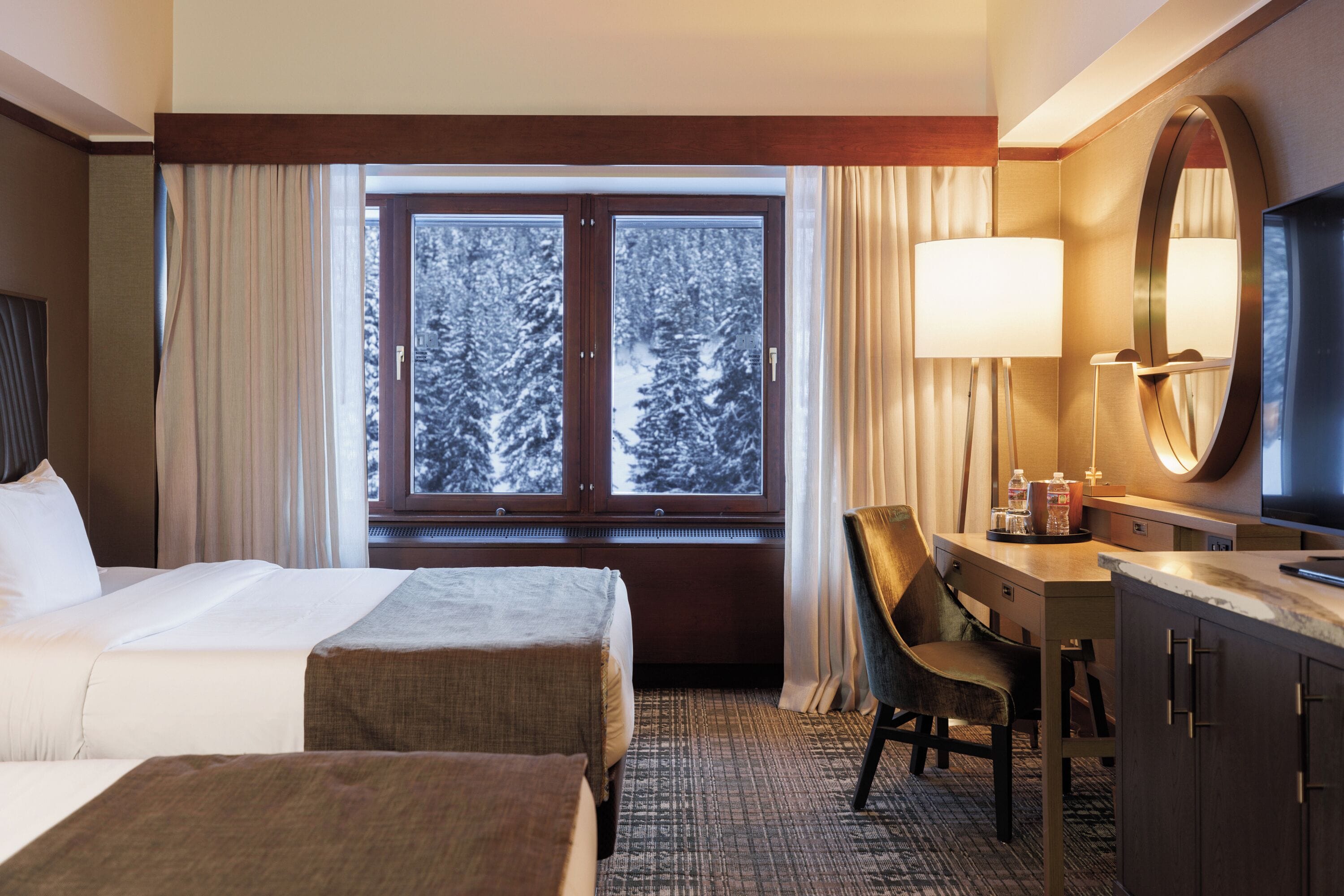 A hotel room with two beds, a desk with a chair, a large window with a view of snow-covered trees, and a mirror on the wall.
