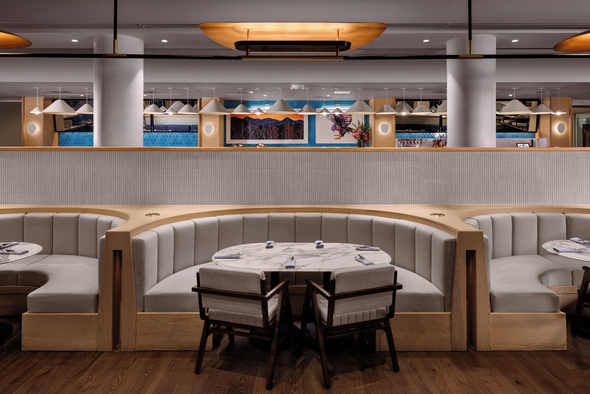 A modern restaurant with a central round table and booths, overhead pendant lighting, and a bar area in the background.