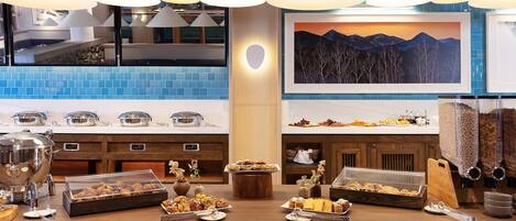 A breakfast buffet with various food items, a painting of a mountain landscape, and a blue-tiled backsplash.