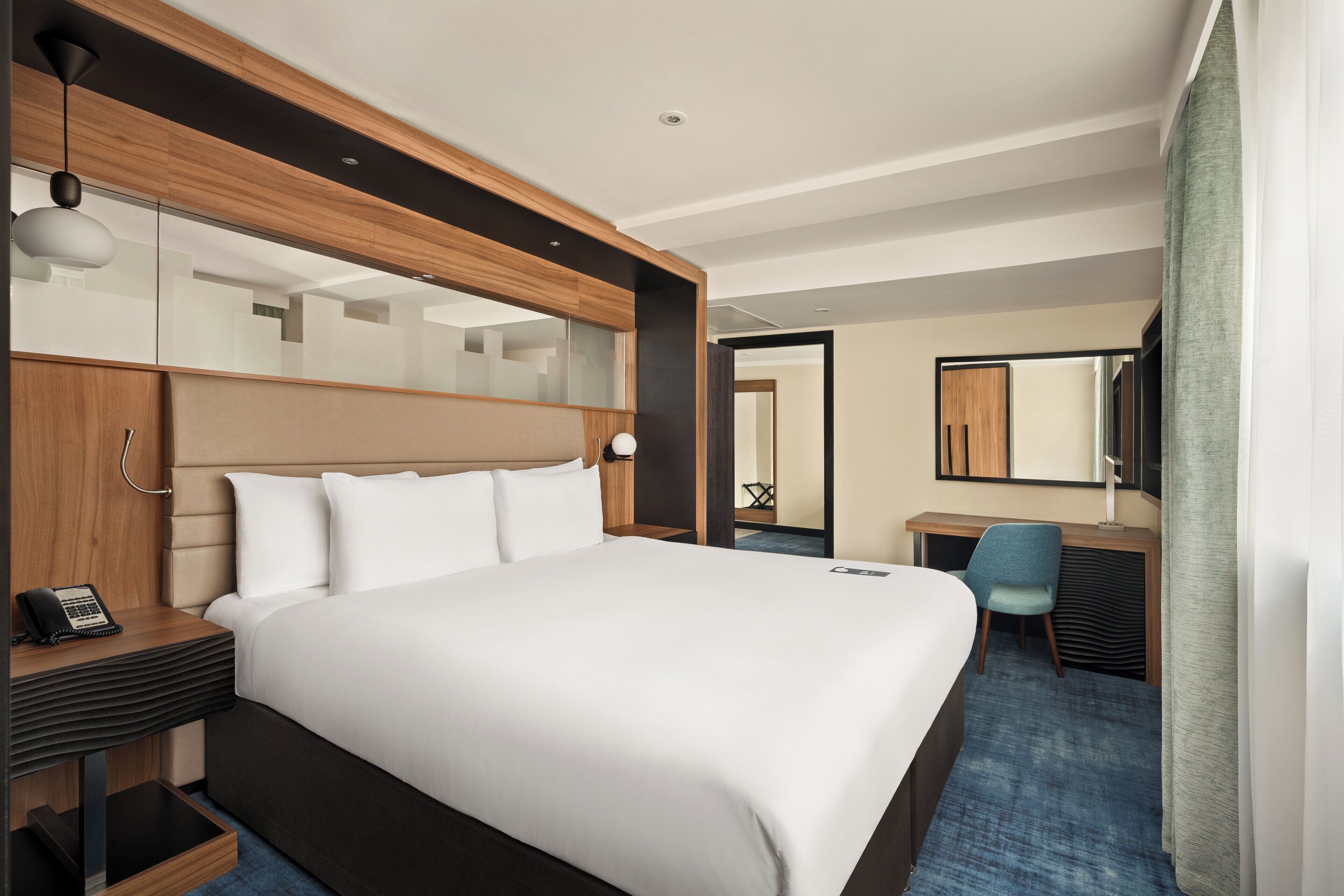 A modern hotel room with a large bed, a desk, a chair, a mirror, and a phone on the nightstand.