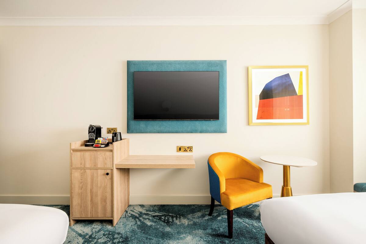 A hotel room with a bed, a desk with a flat-screen TV, a chair, a small table, and a painting on the wall.