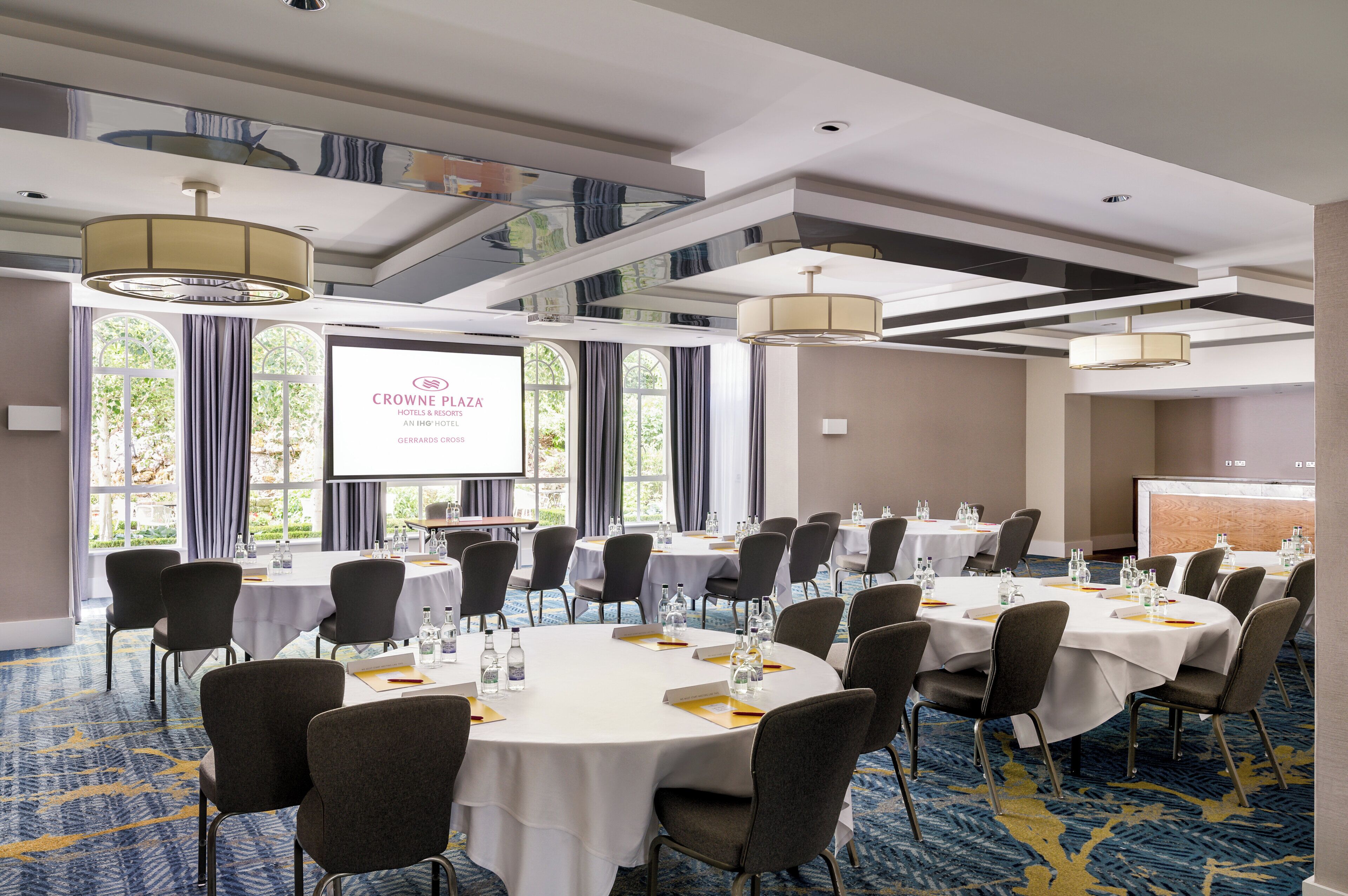 A conference room with multiple tables and chairs, a large screen displaying 'CROWNE PLAZA,' and a view of greenery outside.