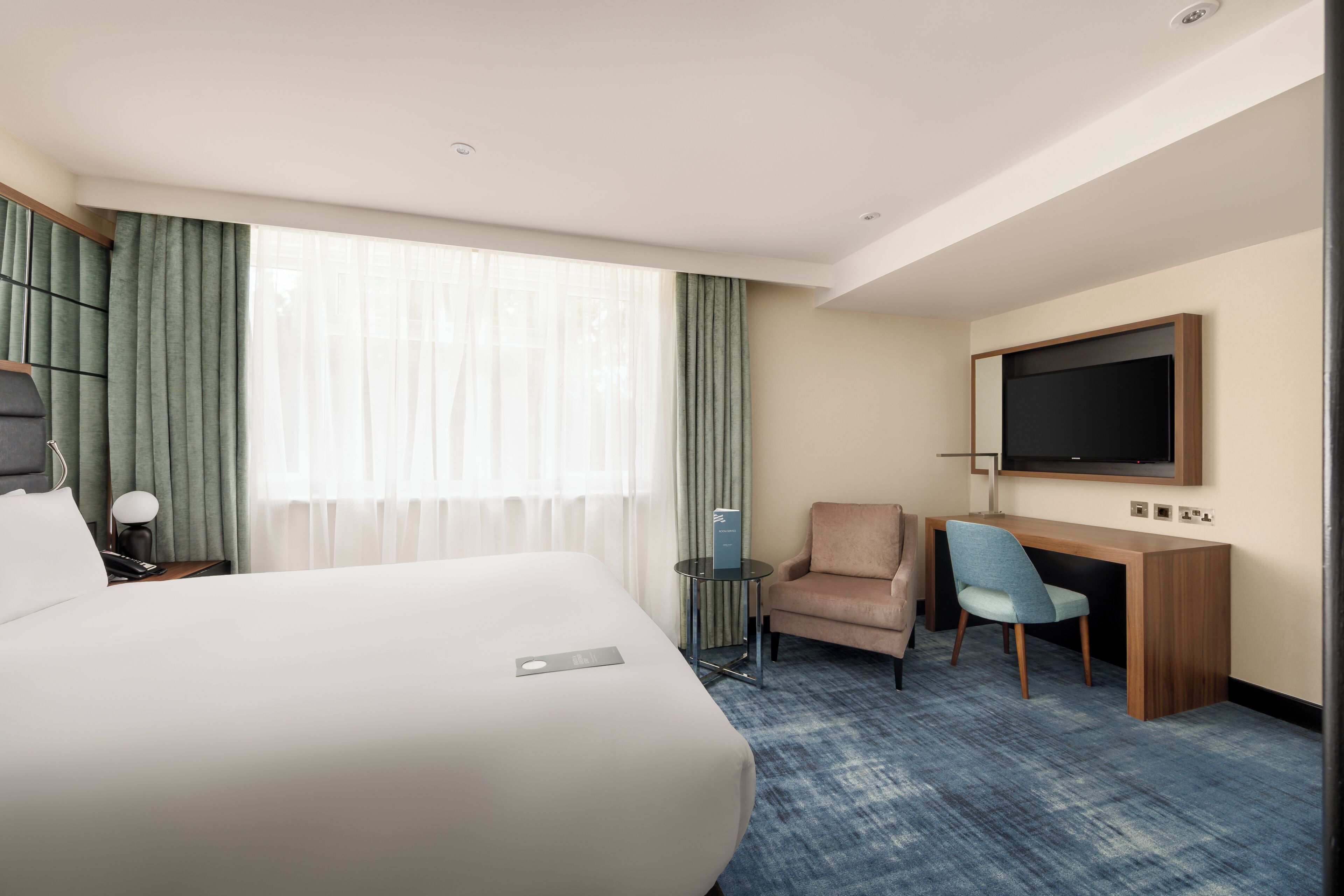 A hotel room with a large bed, a desk with a chair, a TV mounted on the wall, and a window with curtains.