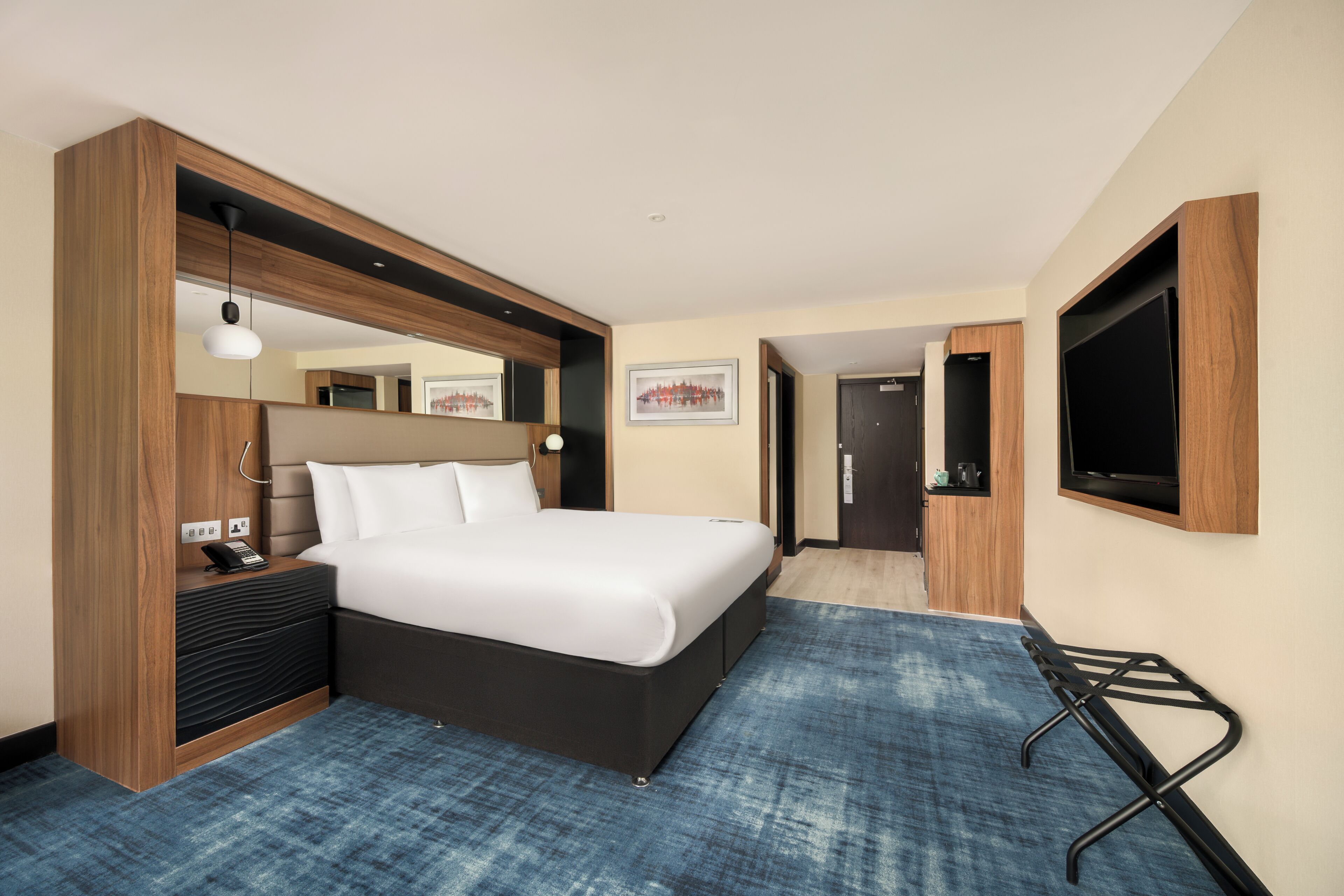 A hotel room with a bed, bedside table, lamp, and a TV mounted on the wall.
