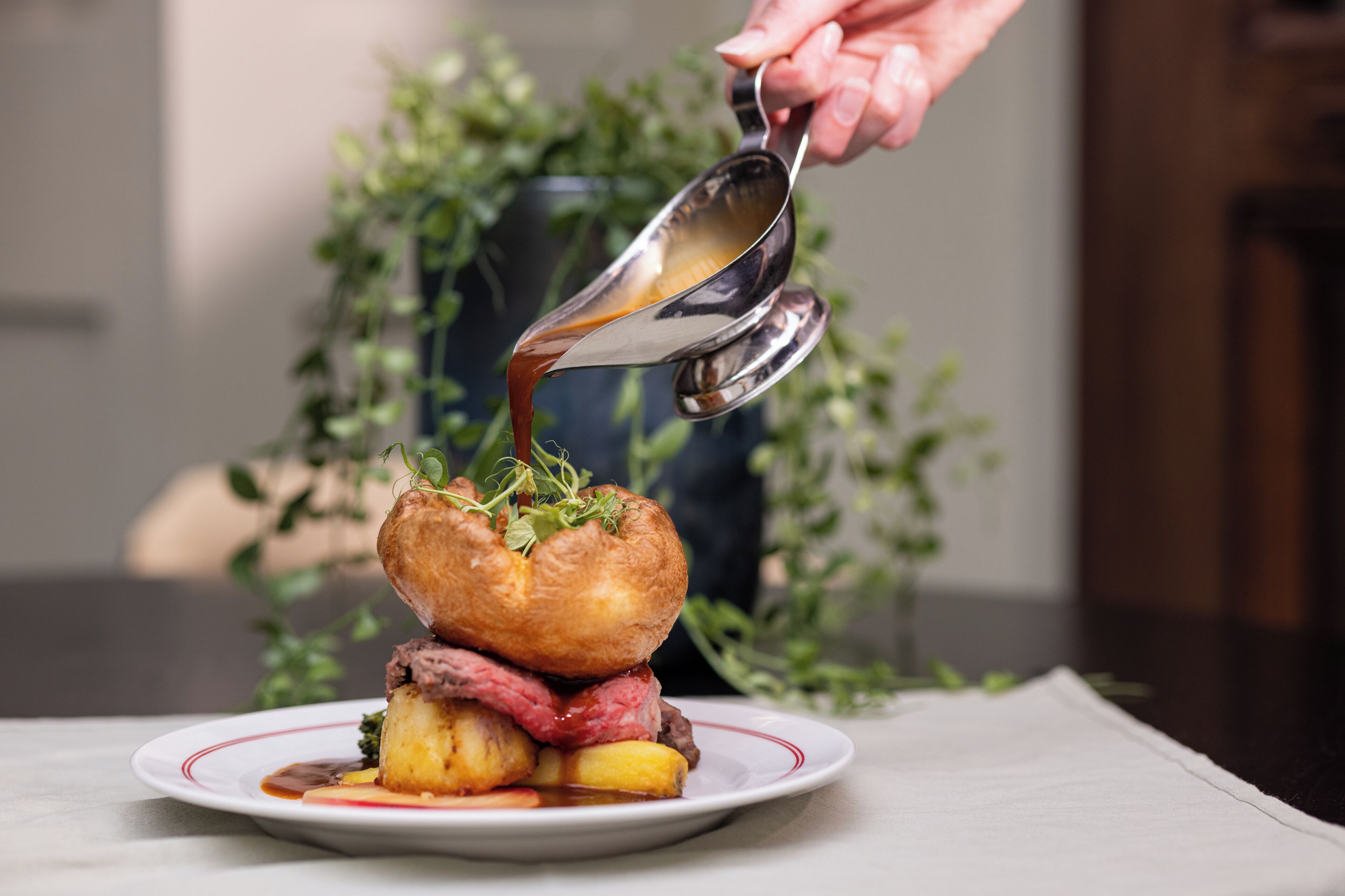 A plate of food with a meat and potato dish, a sauce being poured over it, and a hand holding a spoon.
