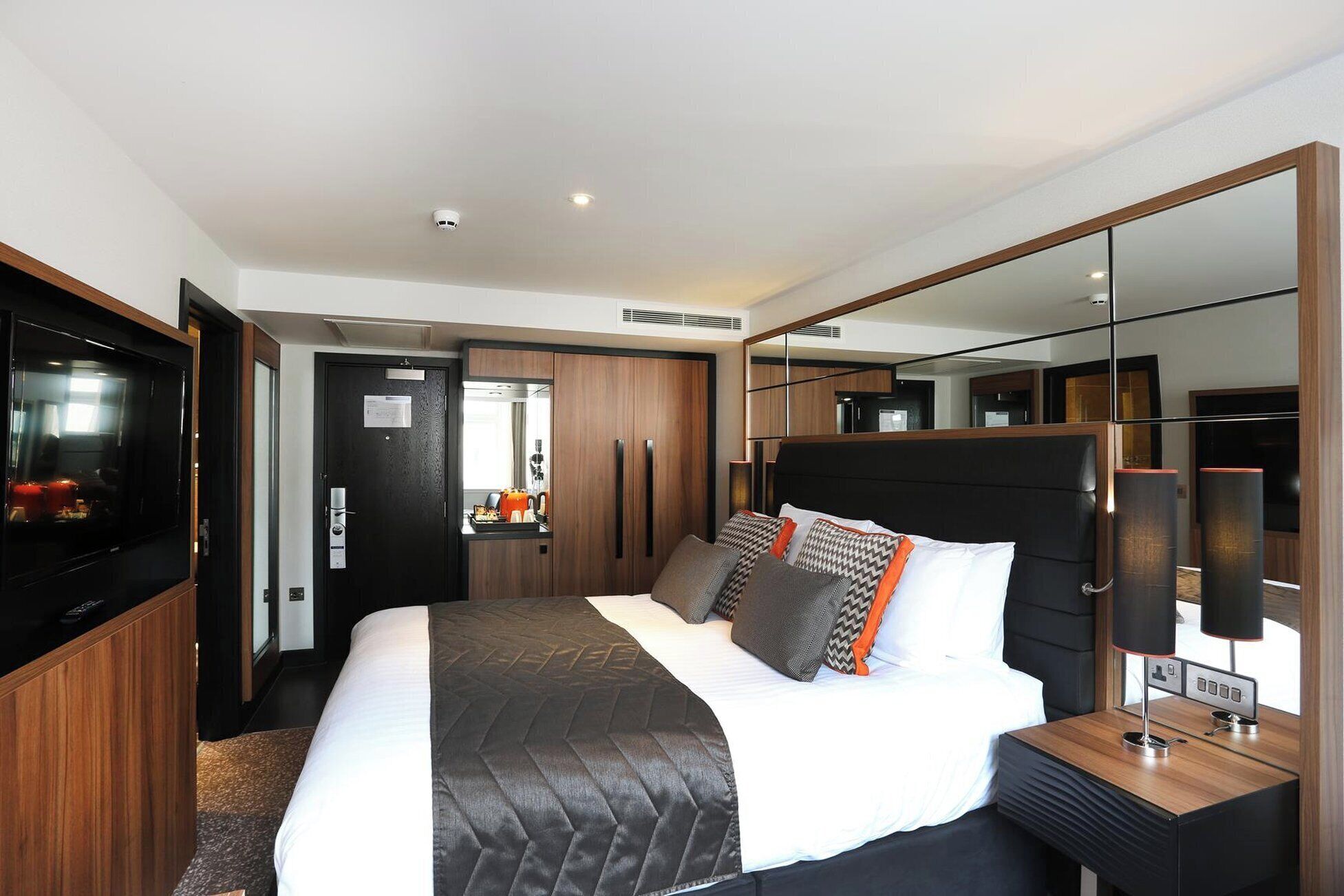 Ein modernes Hotelzimmer mit einem großen Bett, einem Fernseher, einem Spiegel und einem Nachttisch.