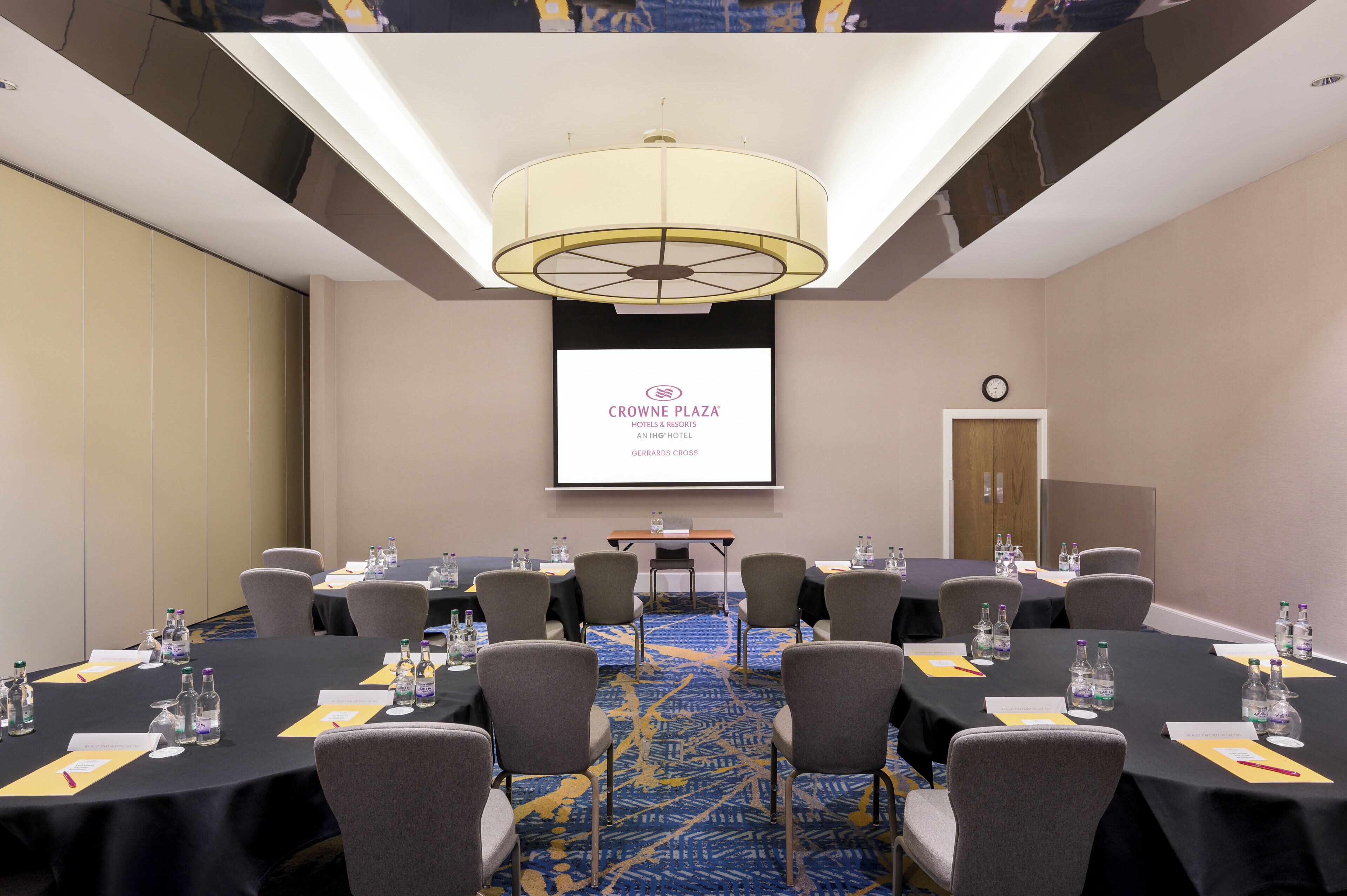 Un salón de conferencias con varias mesas, sillas y una pantalla proyectora mostrando el logotipo de Crowne Plaza.