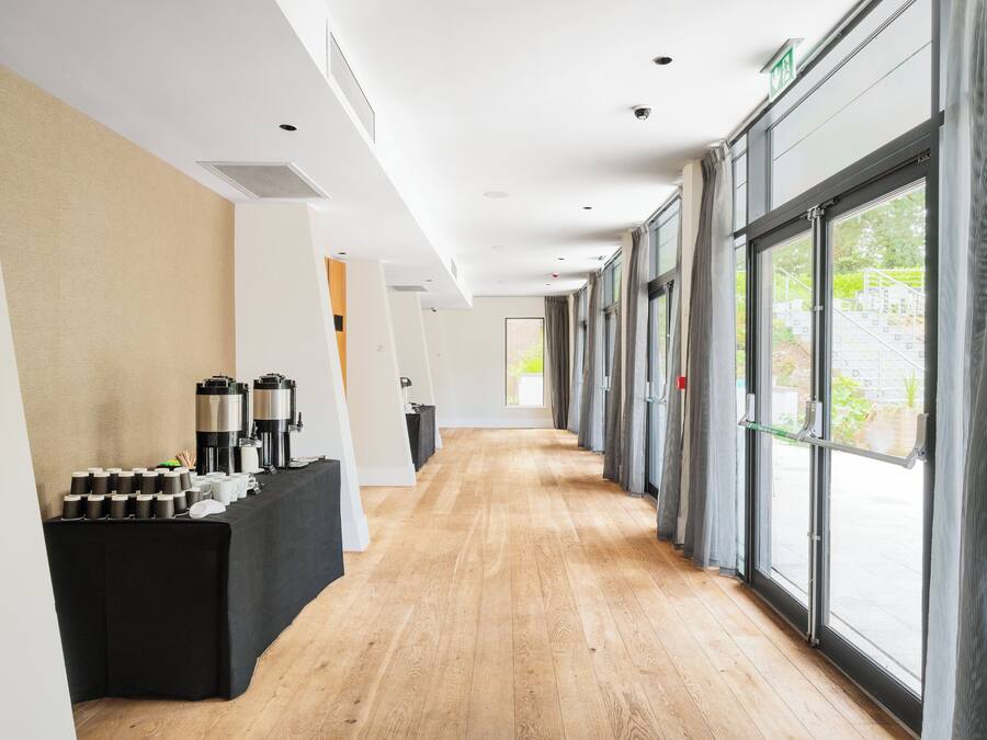 Un pasillo de hotel con una estación de café, puertas numeradas y ventanas amplias.