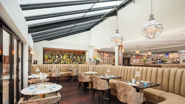 A modern interior with a high ceiling, a seating area with a sofa and chairs, and a bar with plants and framed pictures. - Crowne Plaza Gerrards Cross by IHG (Beaconsfield)