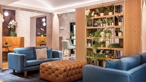 A modern living room with a blue sofa, a brown ottoman, and a wooden shelving unit filled with plants. - Crowne Plaza Gerrards Cross by IHG (Beaconsfield)