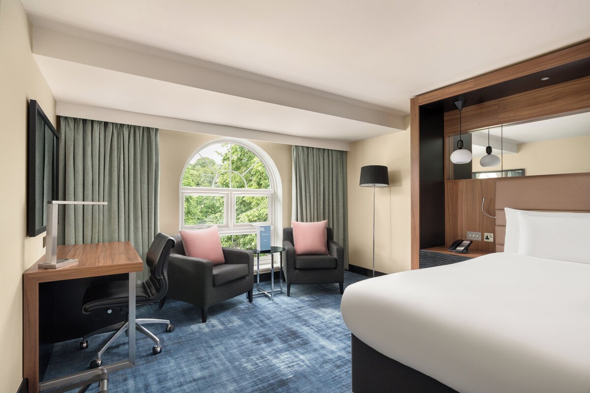 Une chambre d’hôtel comprenant un lit, deux fauteuils, un bureau, une petite table, une lampe, un téléphone et une fenêtre avec des rideaux.