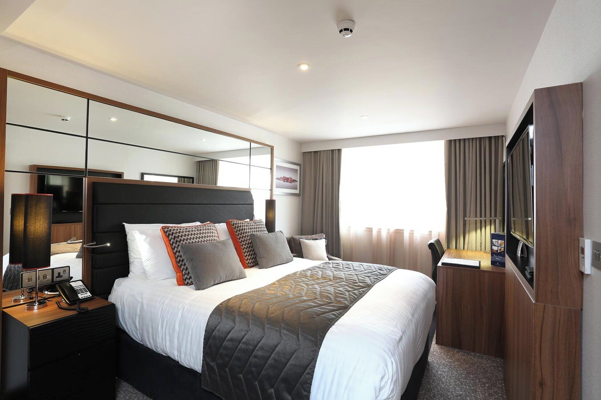 A modern hotel room with a large bed, a bedside table with a phone, a flat-screen TV, and a large mirror.