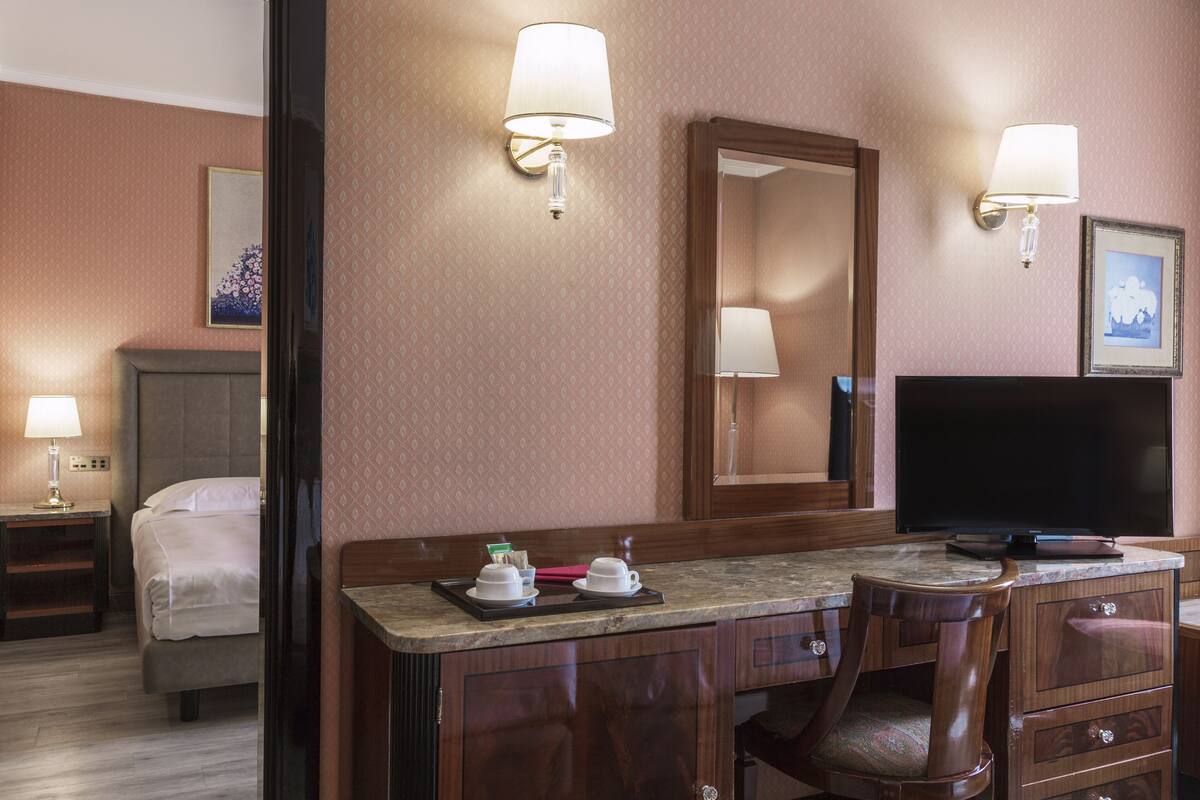 A hotel room with a bed, a desk with a flat-screen TV, a chair, and a mirror.